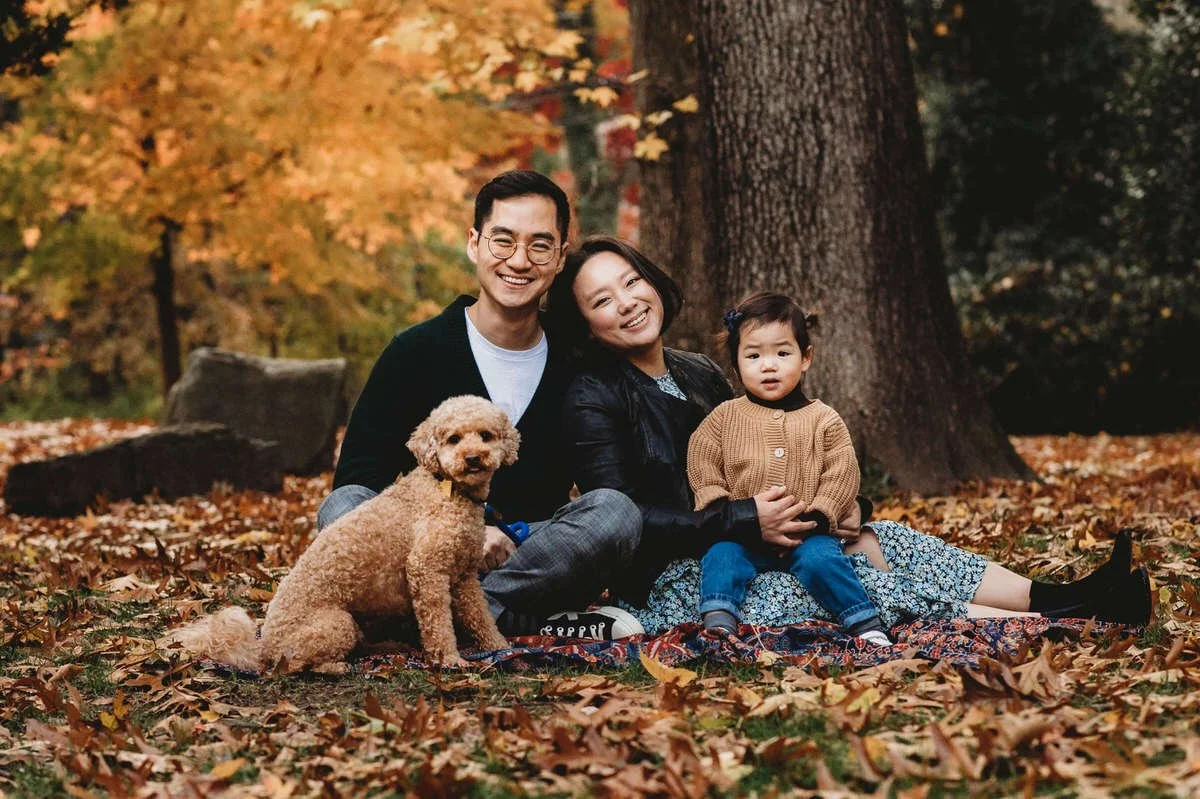 Family of three with a dog sitting on a blanket in an autumn park.