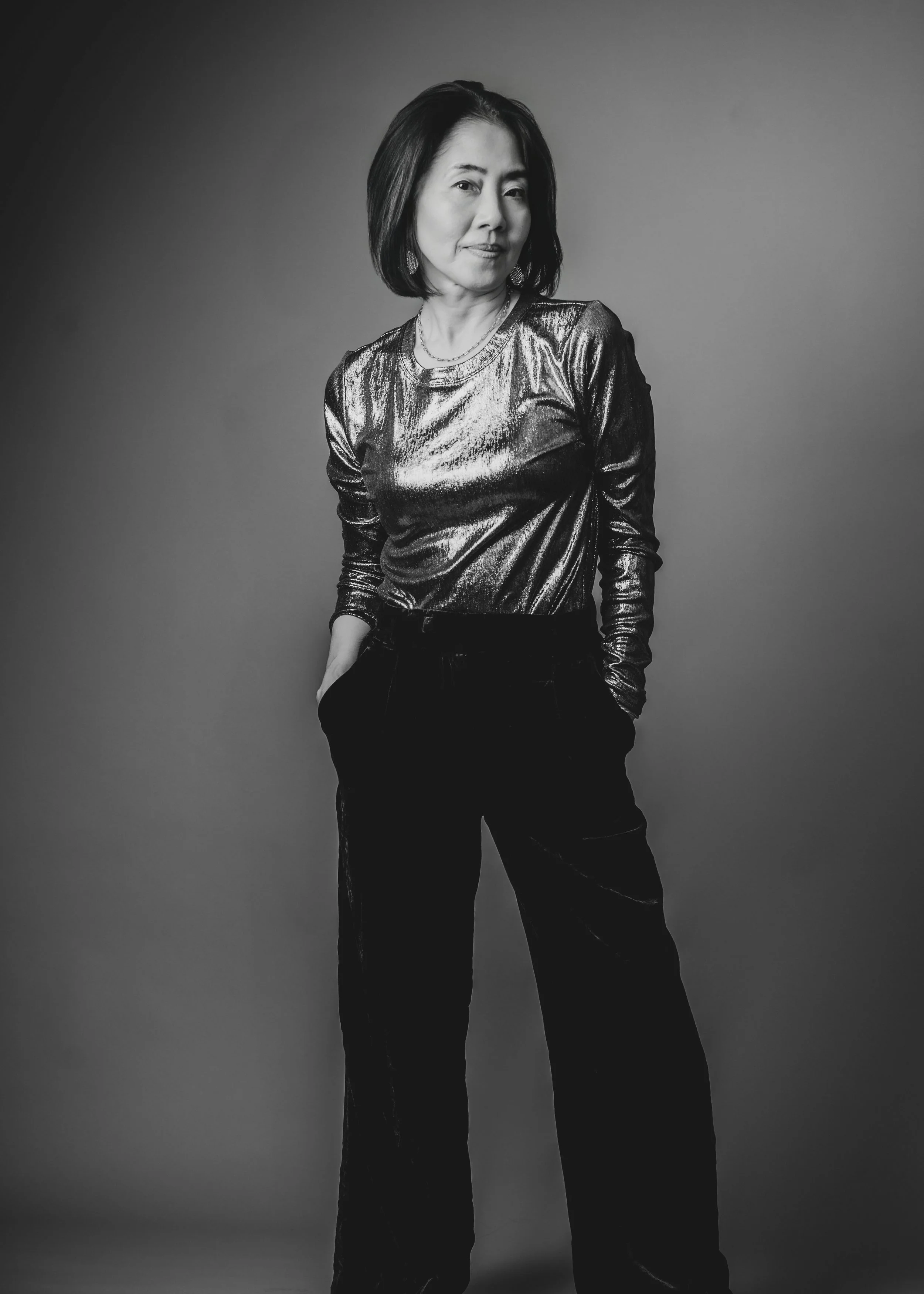 Black and white photo of a woman with shoulder-length dark hair, wearing a shiny metallic long-sleeve top and dark pants, standing with hands in her pockets against a plain background.