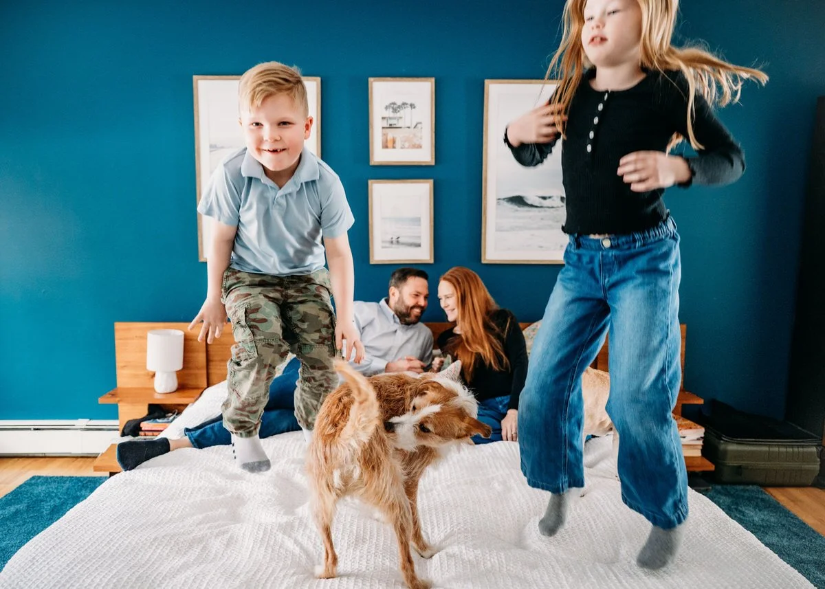 Children jumping on a bed while a dog stands in the center, with a couple sitting on the bed in the background.