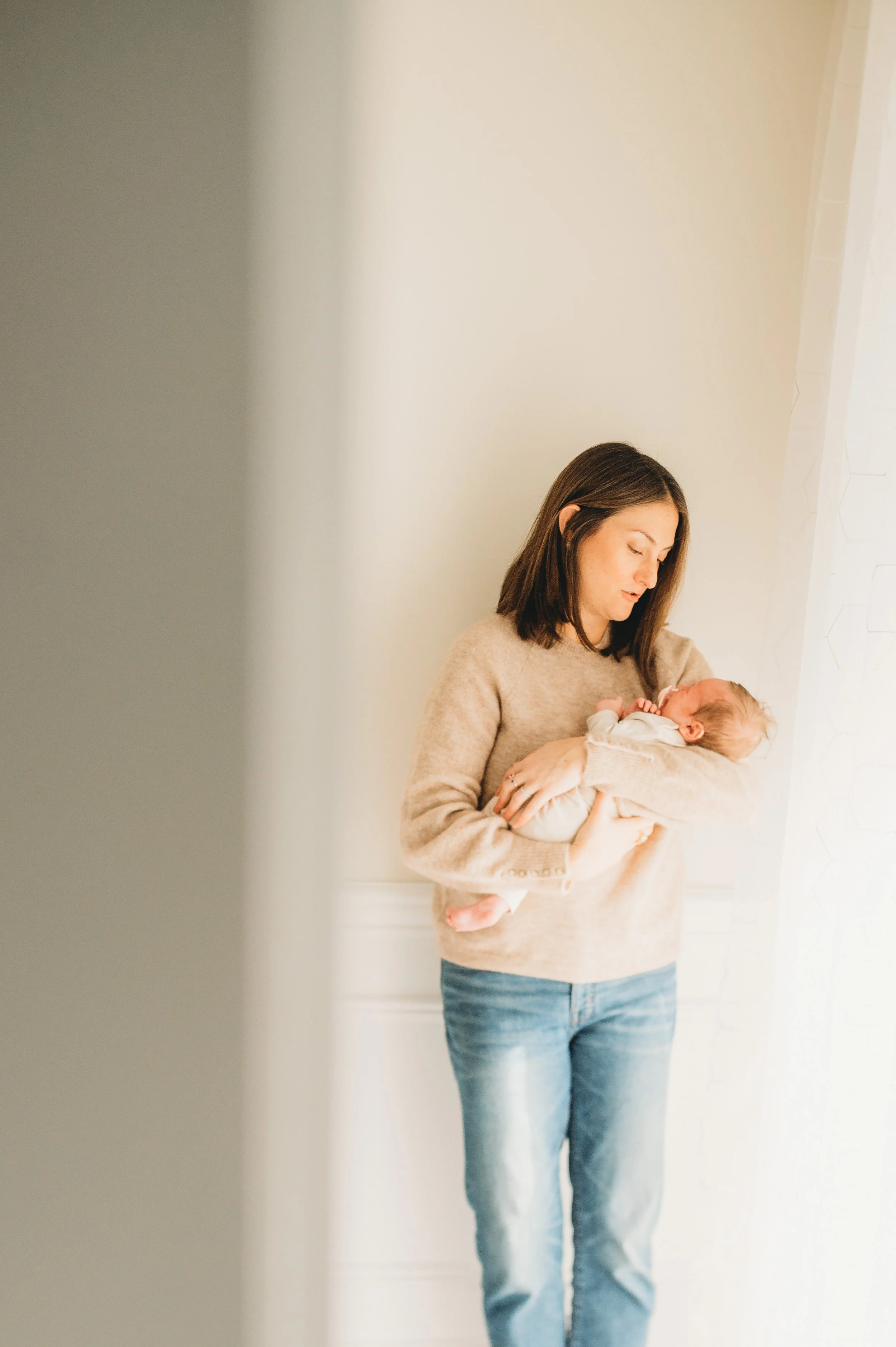 A woman wearing a beige sweater and jeans is holding a newborn baby in her arms while standing near a window with white curtains.