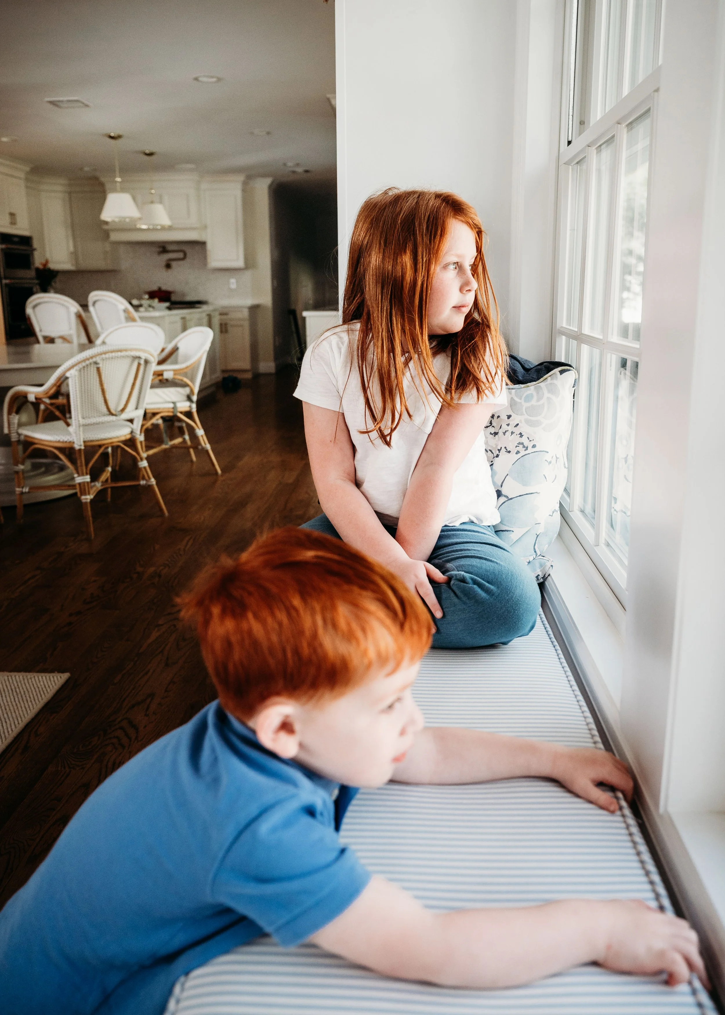 A young girl with red hair watching outside through a window while sitting on a cushioned window seat, and a young boy with red hair lying on the floor also looking outside.