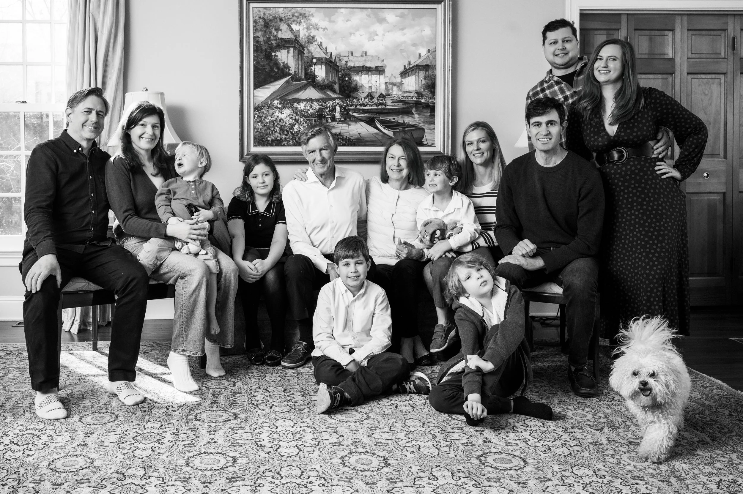 A large multi-generational family posing together in a living room. There are 13 people and one dog. The family includes children, teenagers, adults, and elderly members. They are sitting and standing in front of a framed painting, with natural light coming from windows on the left.