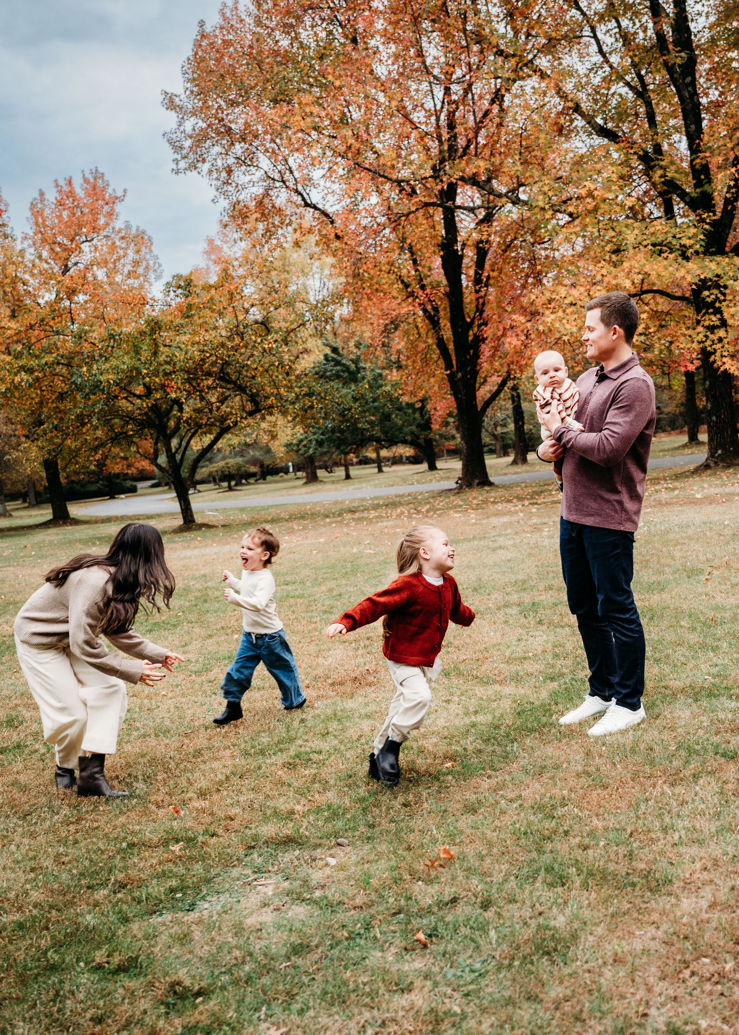 A man holding a baby and four children playing and running on grass in a park during fall, with trees showing colorful autumn leaves in the background.