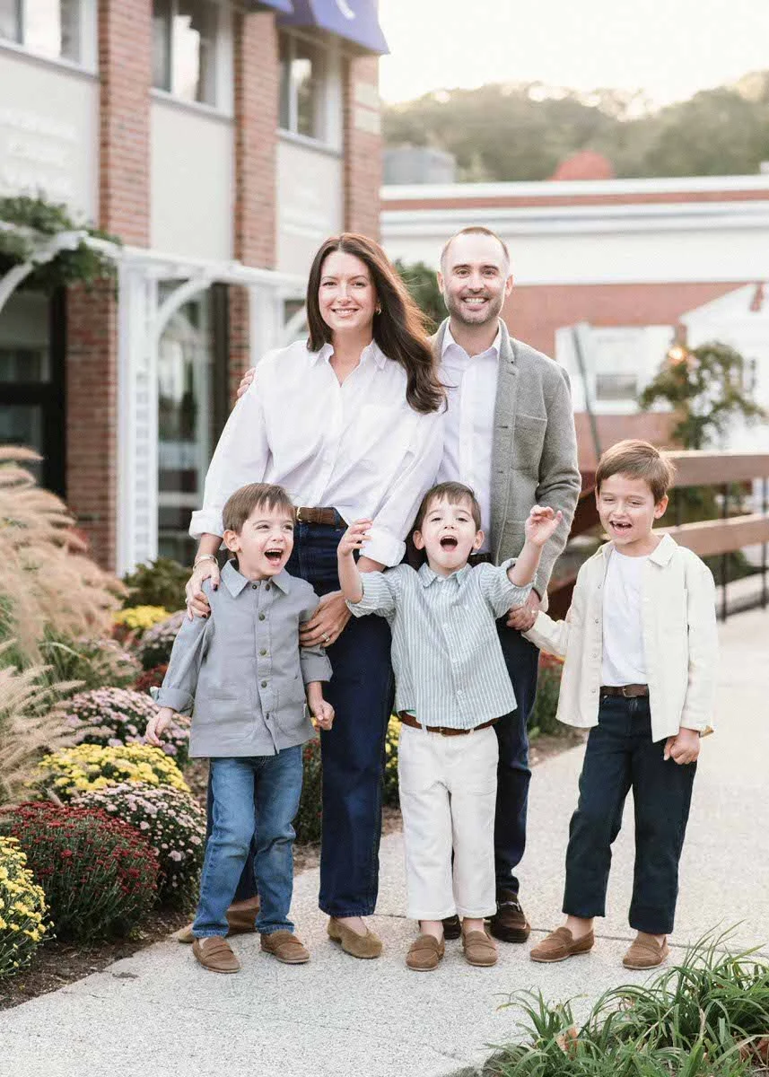 A happy family of five, including a mother, father, and three young boys, standing outdoors on a sidewalk with a building and landscaping in the background, enjoying a sunny day.