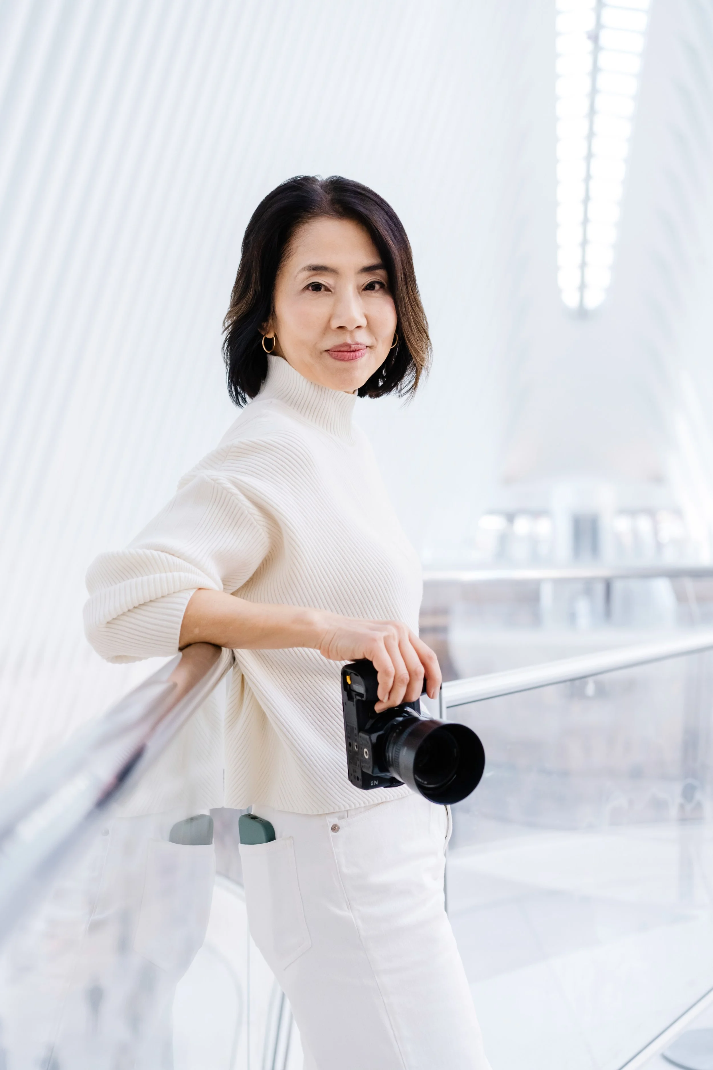 A woman with shoulder-length dark hair wearing a white turtleneck sweater and white pants holds a camera while standing in a modern, white, curvilinear architectural space.