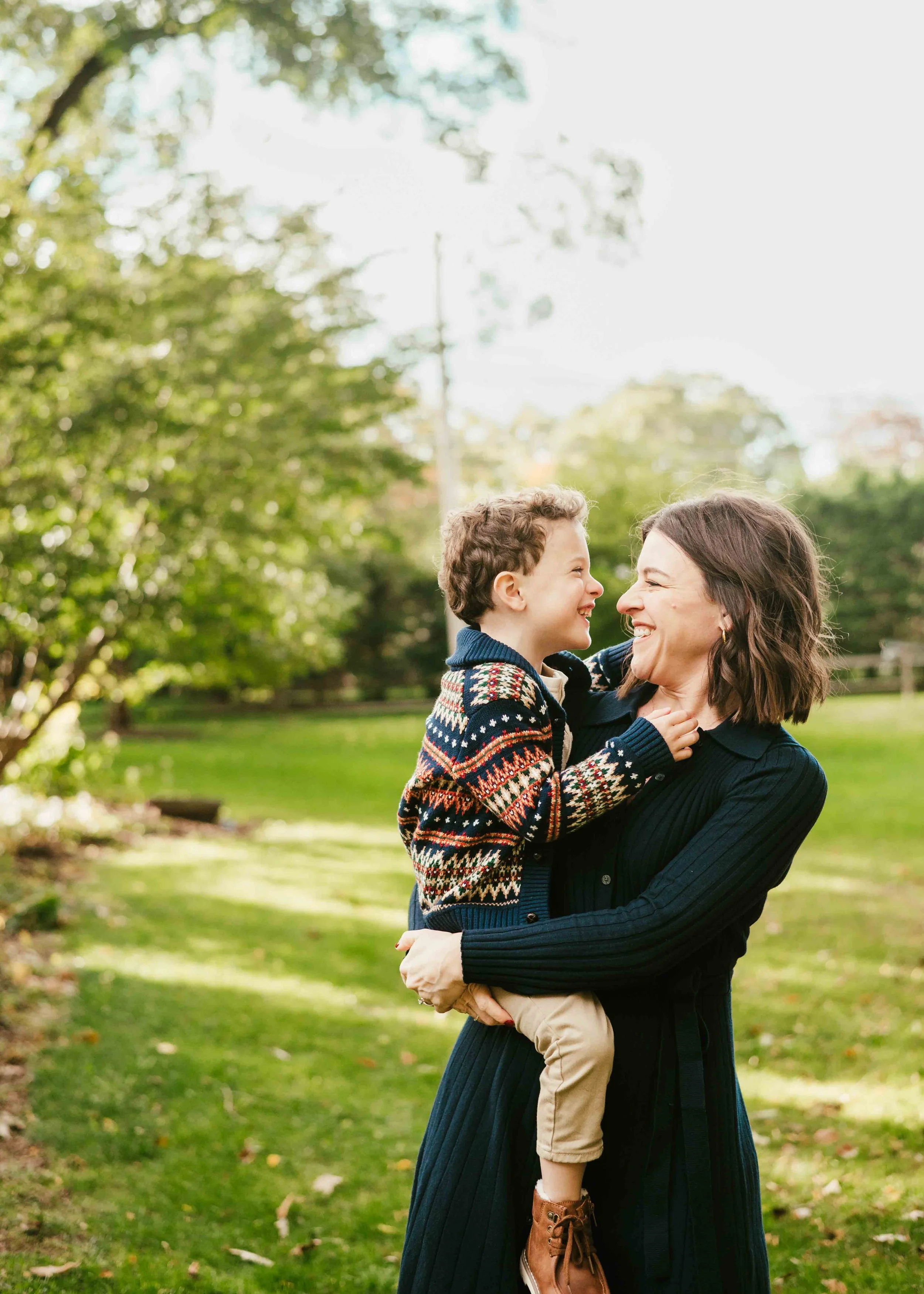 A woman holding a smiling boy in an outdoor park setting with trees and grass.