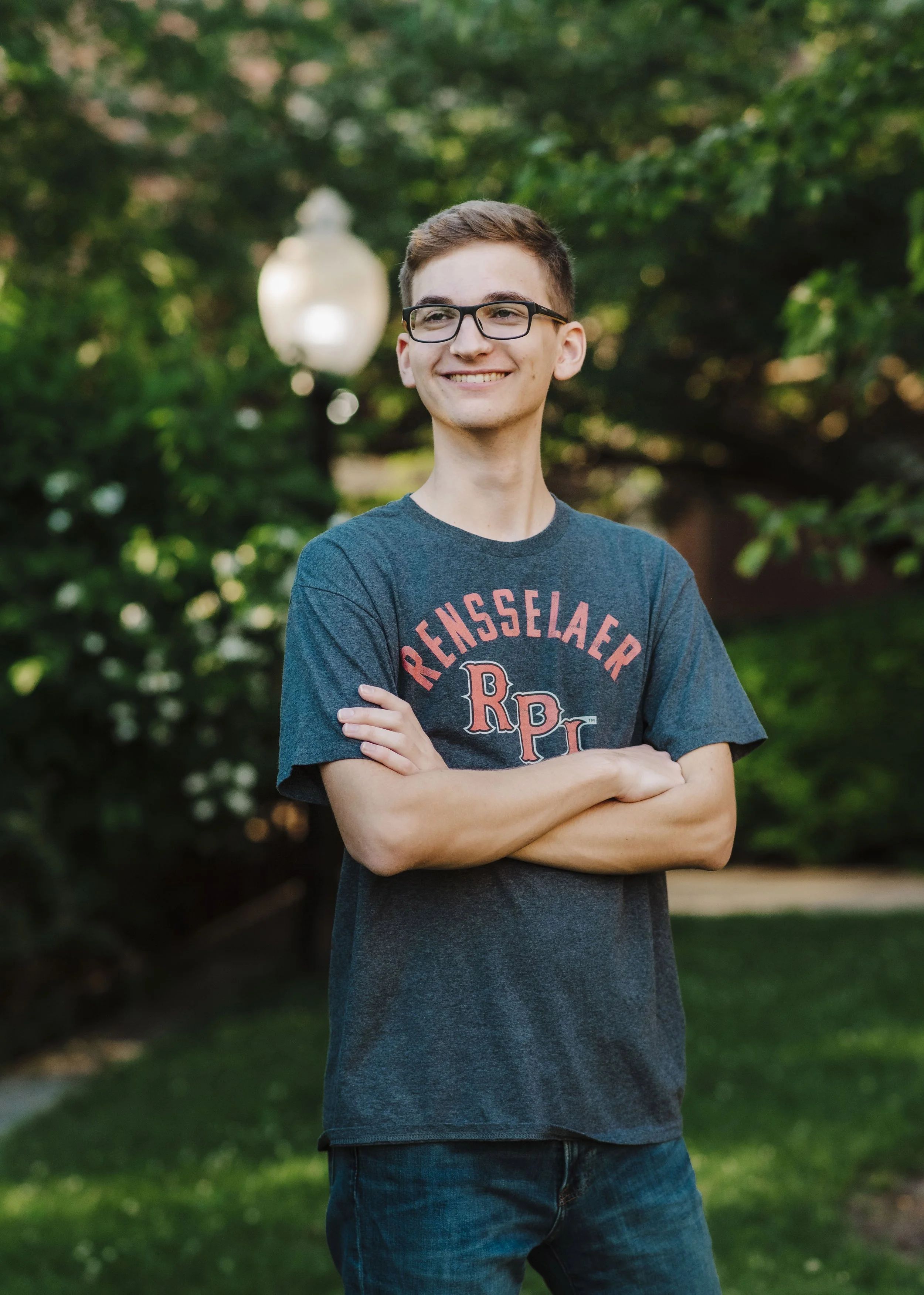 A young man with short brown hair, glasses, and a smile, standing outdoors with arms crossed in front of a green leafy background and a blurred white lamp post.