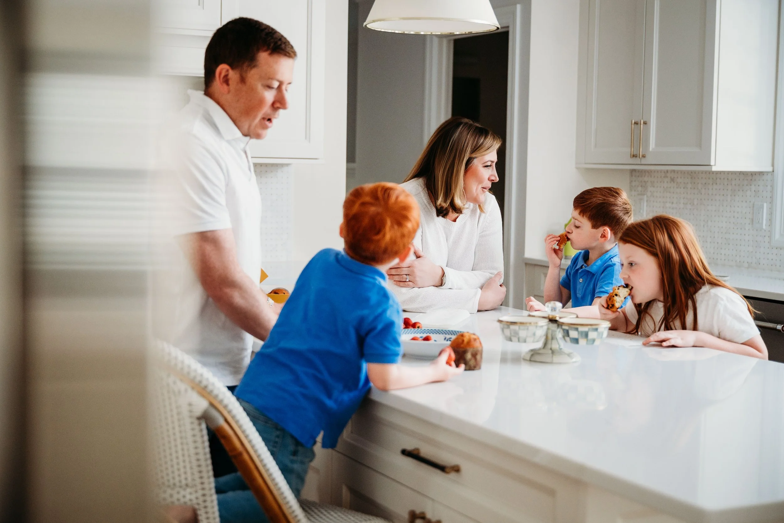 A family of five having breakfast in a bright kitchen. A woman and four children are sitting and standing at the white kitchen island, eating and talking.