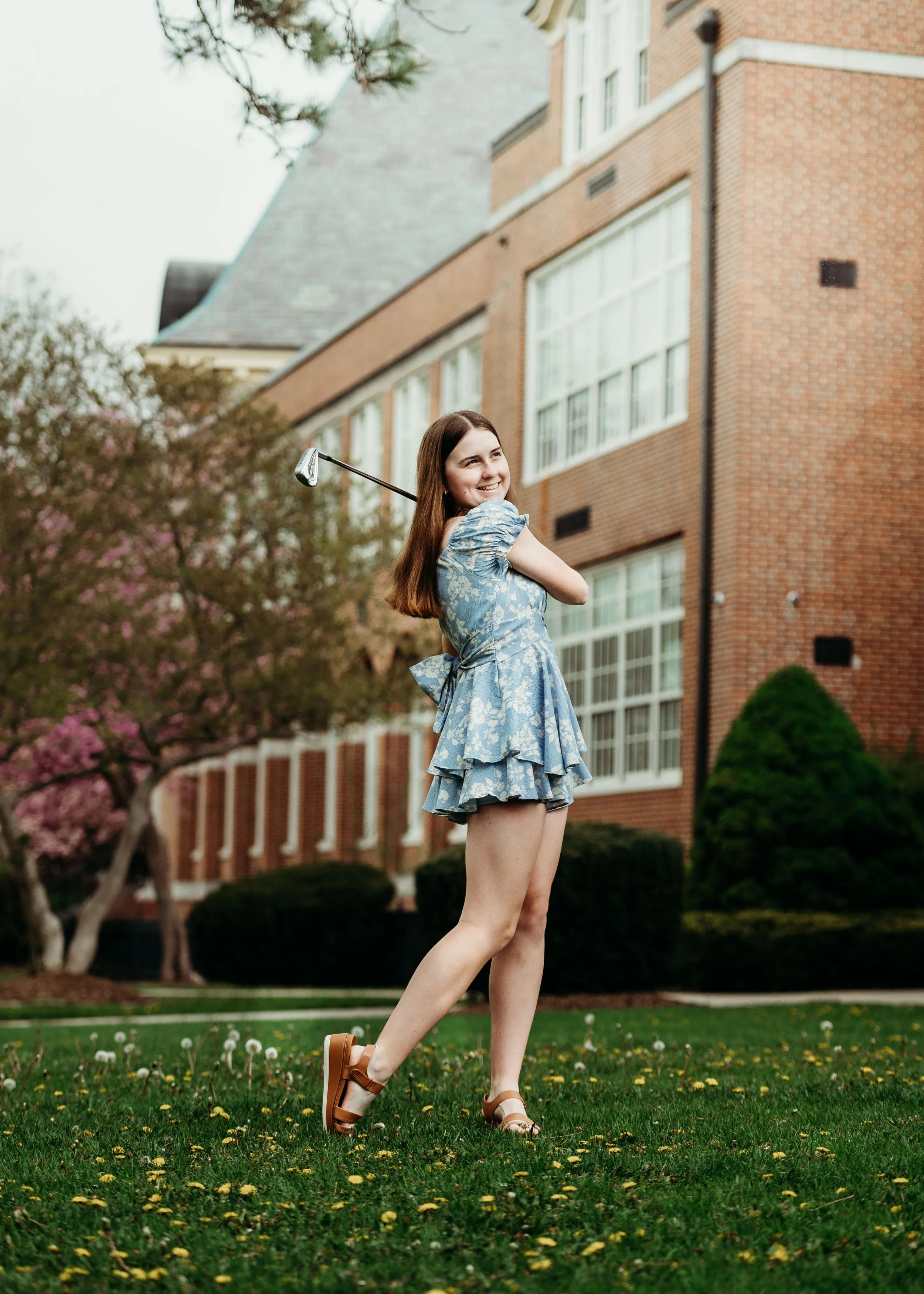 A young woman with long brown hair smiling and holding a golf club on her shoulders, standing on a grassy area with yellow flowers, in front of a red brick building and flowering trees.