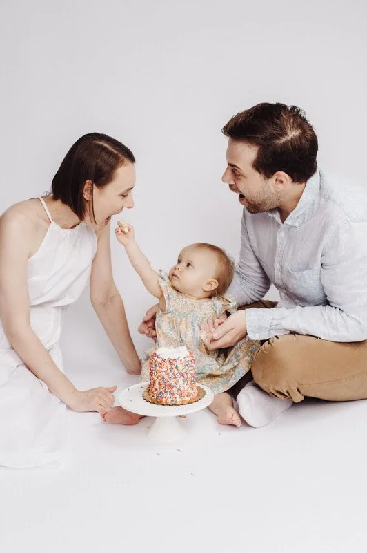 A family celebrating a child's birthday with a small cake with colorful sprinkles, a woman, a man, and a baby girl sitting on the floor.