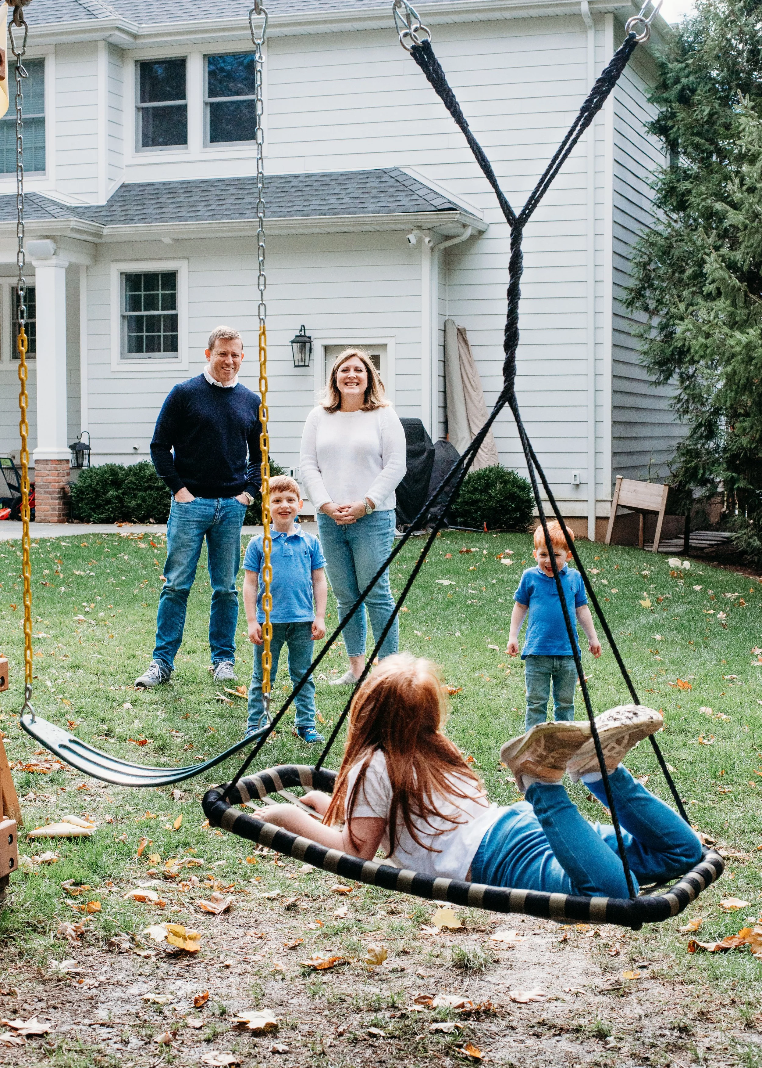 A family enjoys a backyard gathering with children playing on a swing set and parents watching on a grassy lawn, with a house in the background.