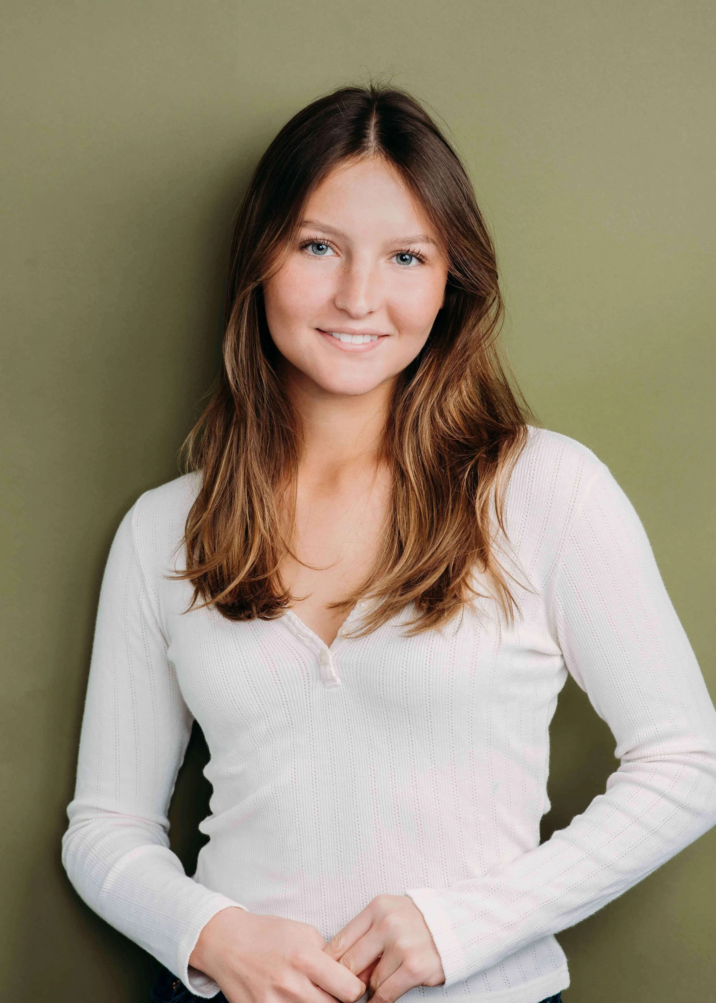Portrait of a young woman with long light brown hair, blue eyes, smiling, wearing a white long-sleeve shirt, standing against an olive green background.