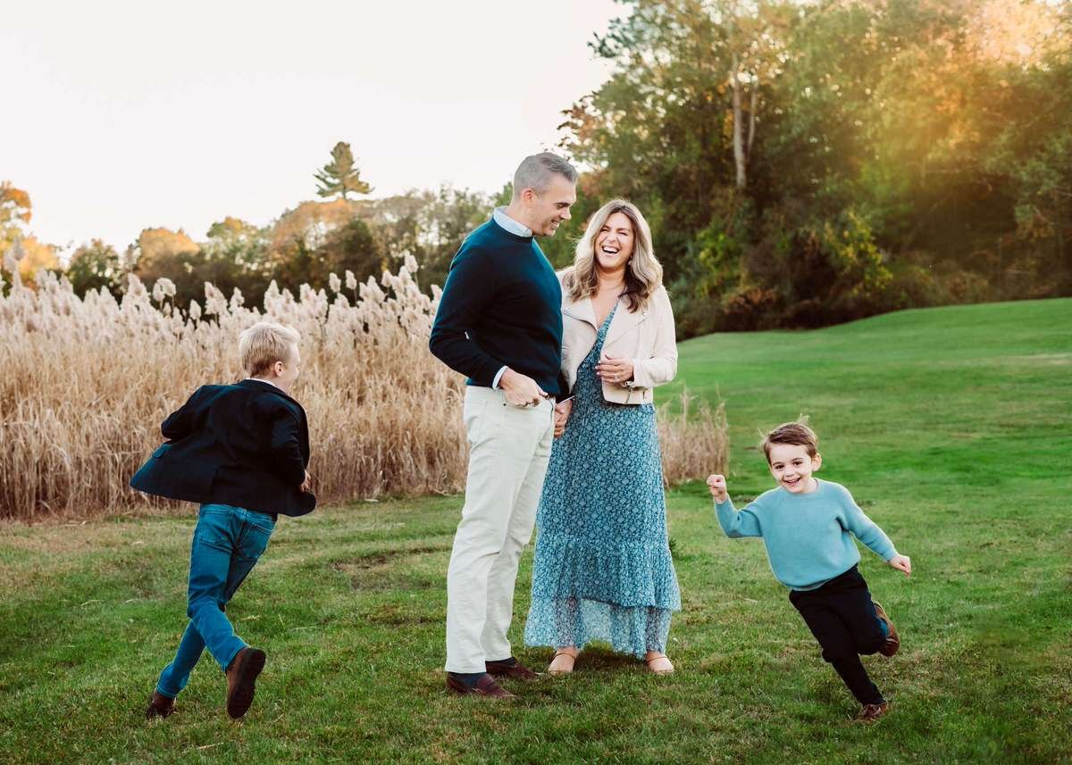 A family of four enjoying outdoors in a park with tall grass and trees, sunset in the background, two children and two adults are smiling and playing.