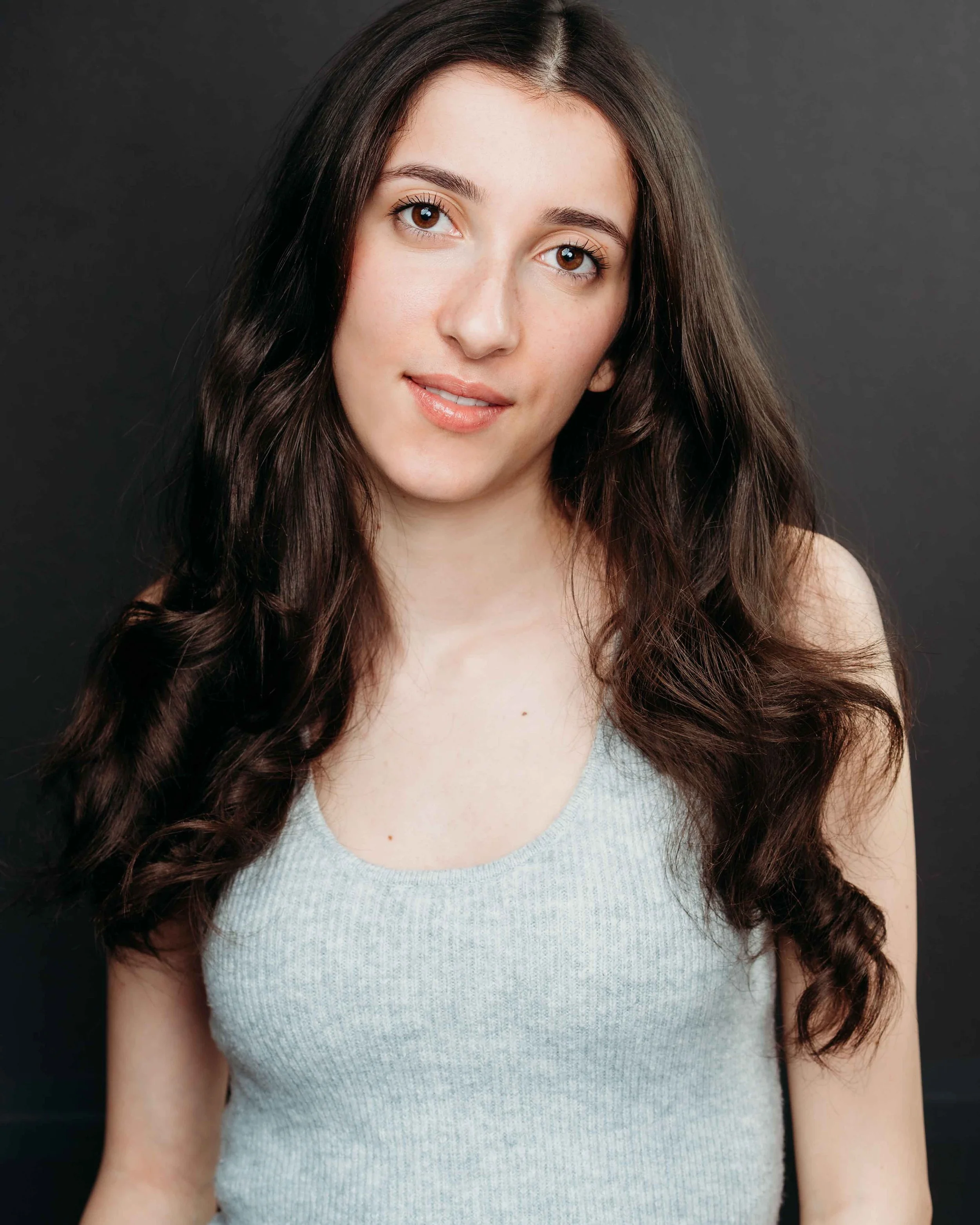 Portrait of a young woman with long, wavy dark hair, wearing a sleeveless gray top, against a plain dark background.
