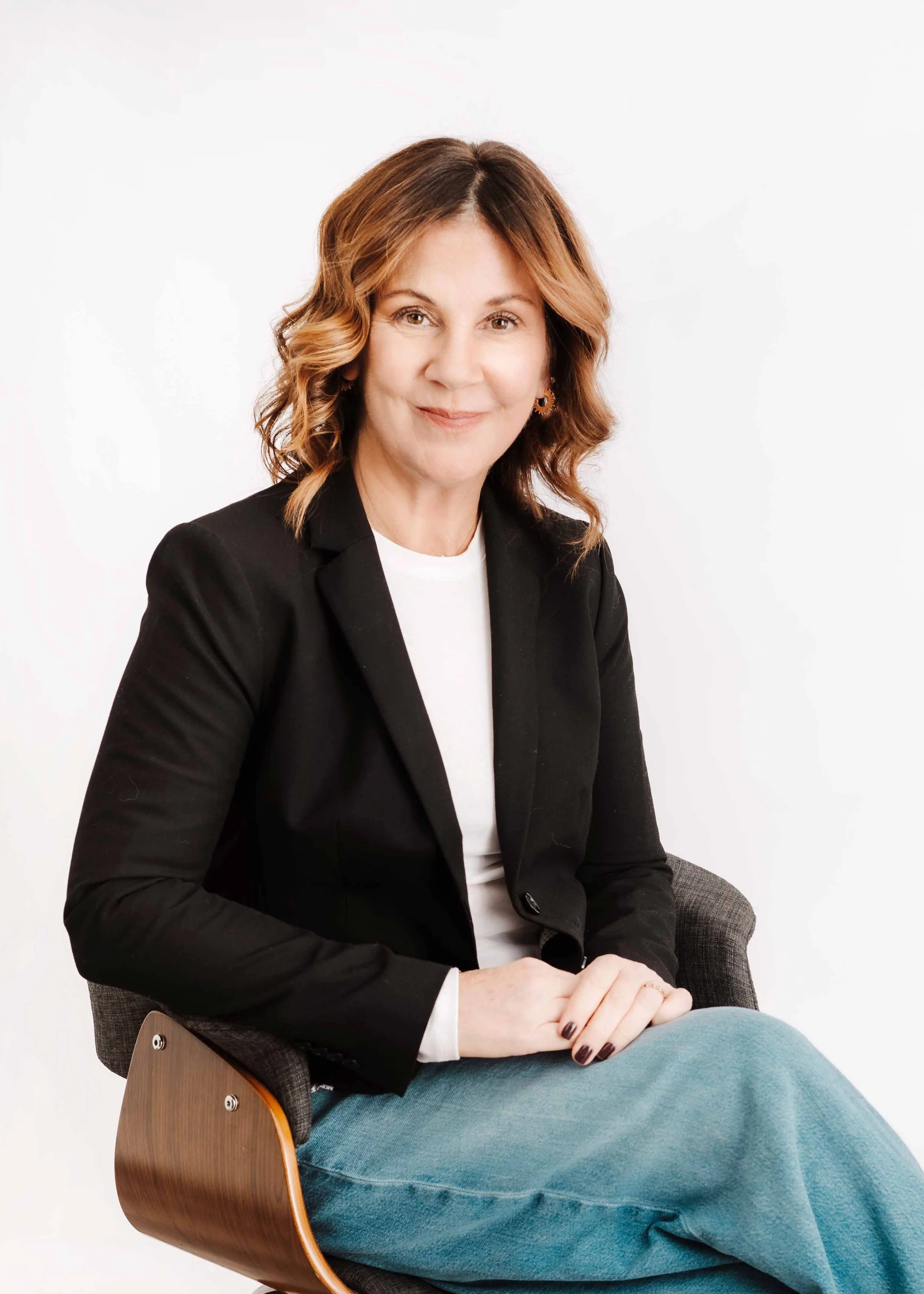 A woman with shoulder-length wavy brown hair and light skin, wearing a black blazer over a white shirt, sitting in a modern chair against a plain white background, smiling at the camera.