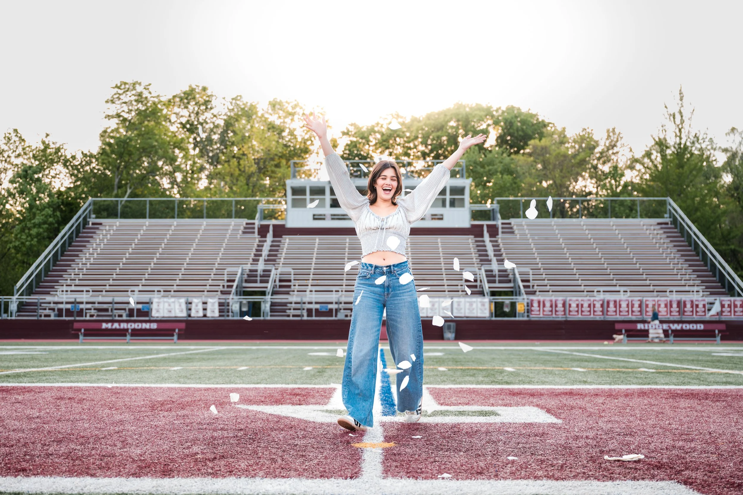 A young woman standing on a football field with her arms raised, smiling, with fall leaves falling around her, and stadium seating in the background.