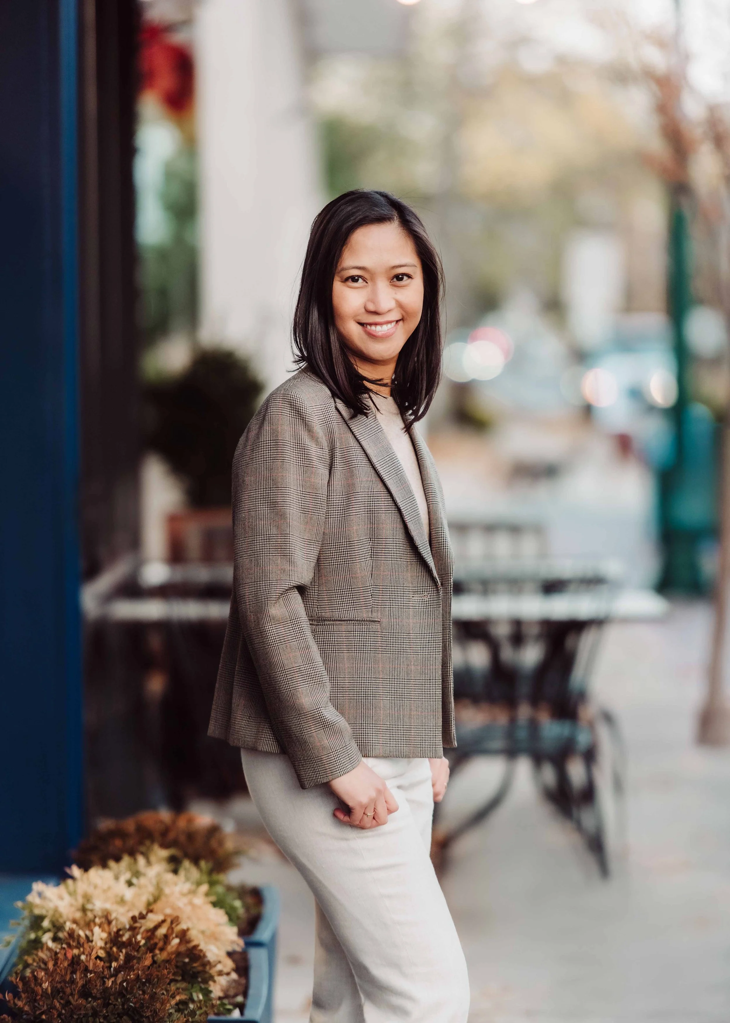 A woman standing outdoors on a sidewalk, smiling, wearing a plaid blazer and light-colored pants, with blurred trees and cars in the background.