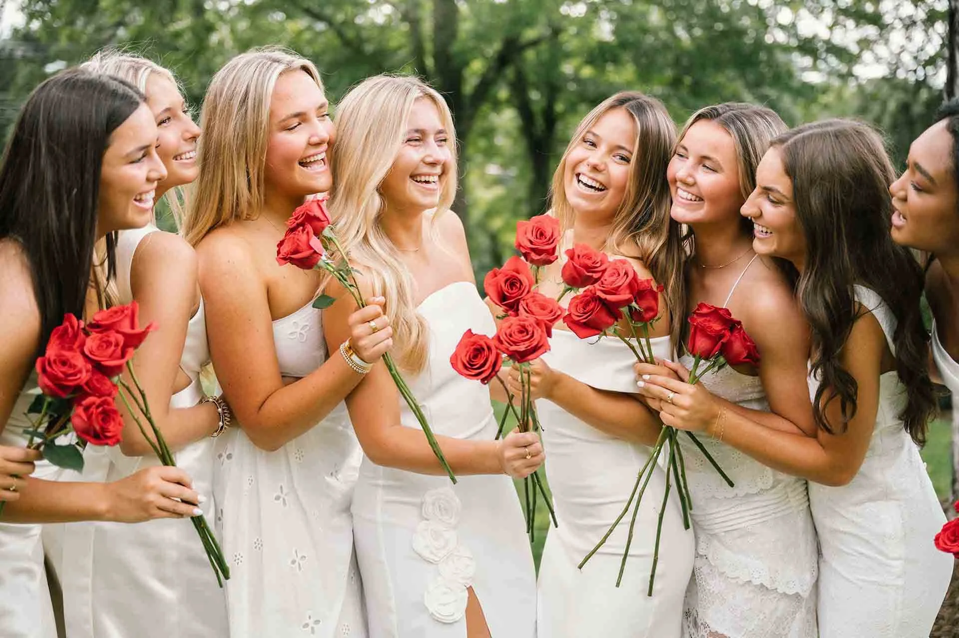 Group of women in white dresses holding red roses, smiling and laughing outdoors in a park.