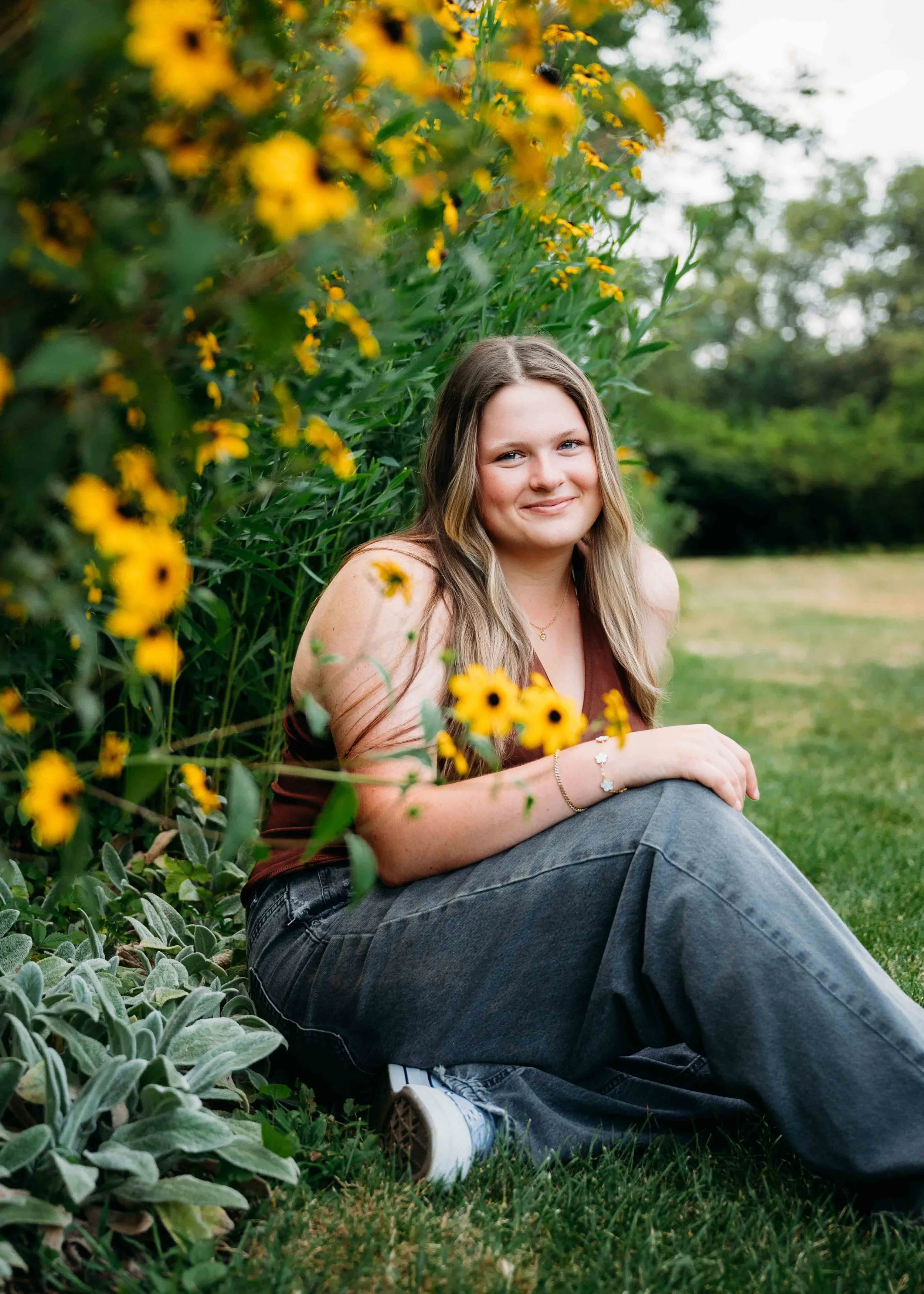 A young woman sitting on grass beside yellow flowers, with green trees in the background, smiling at the camera.