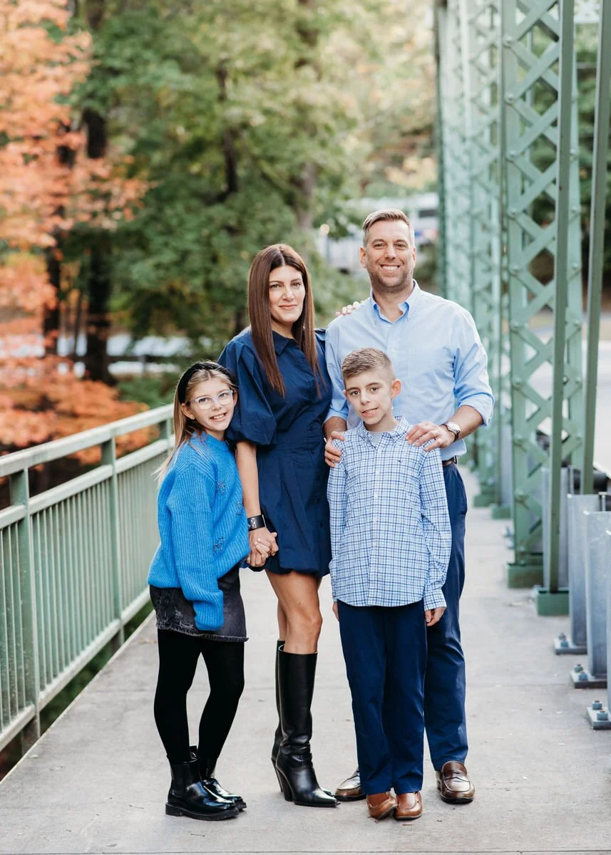 A family of four standing on a bridge with green railing, trees with autumn foliage in the background, and a metal structure on the right side. The family includes a woman dressed in a dark blue dress and black knee-high boots, a man in a light blue 