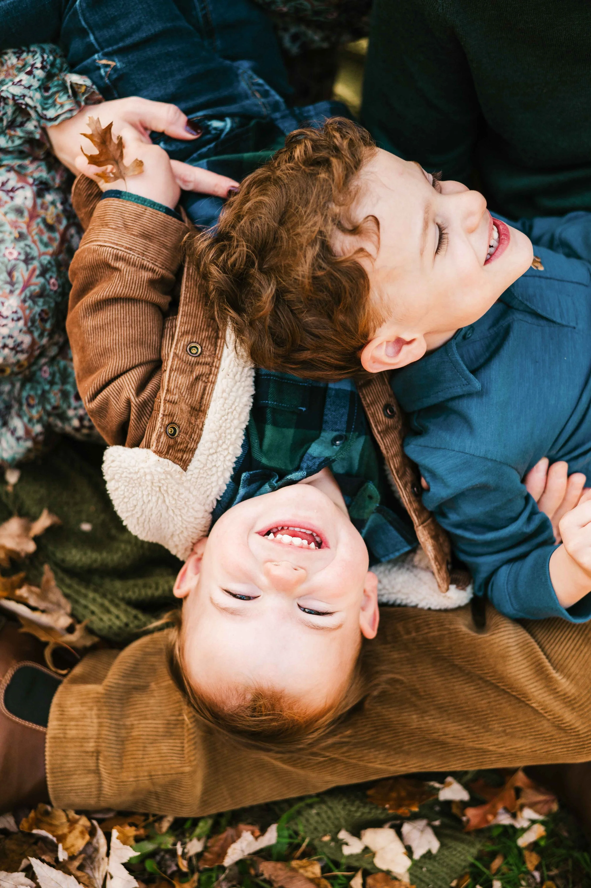 Two young boys are laughing and playing on a bed of fallen autumn leaves outdoors.