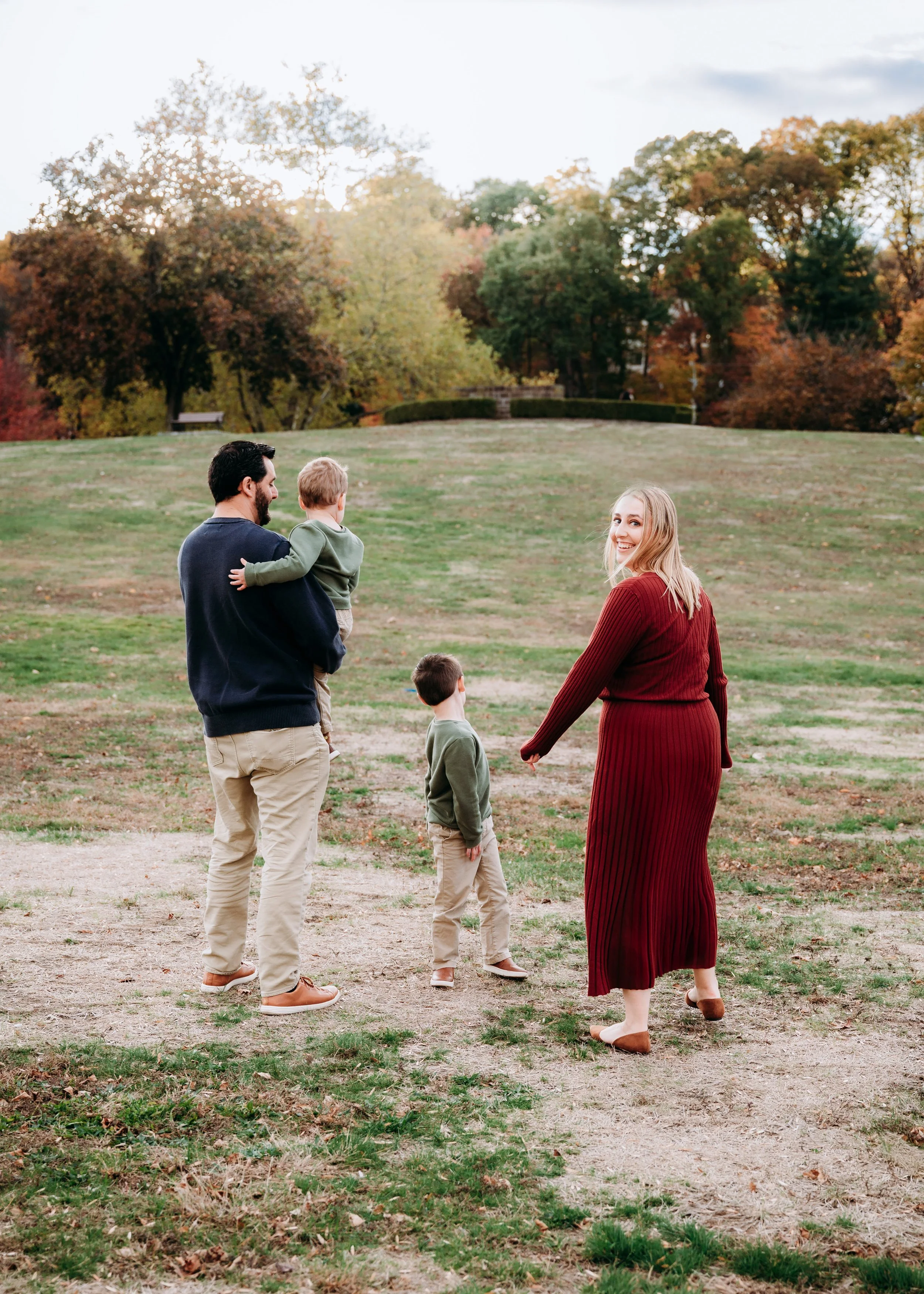 A family of four with two young children, walking in a park during autumn, with colorful trees in the background.
