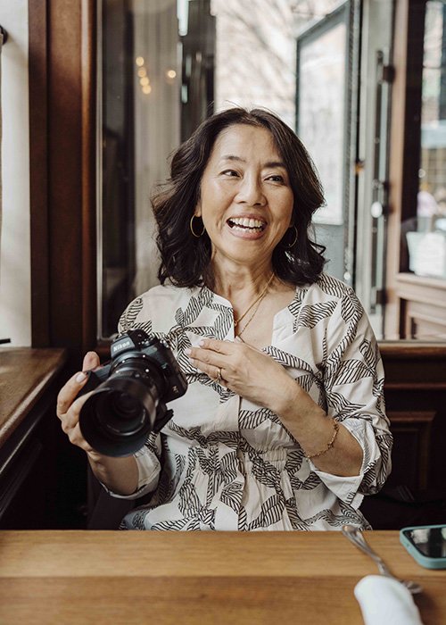 Woman with dark hair smiling and holding a camera, sitting at a wooden table in a cafe or restaurant.