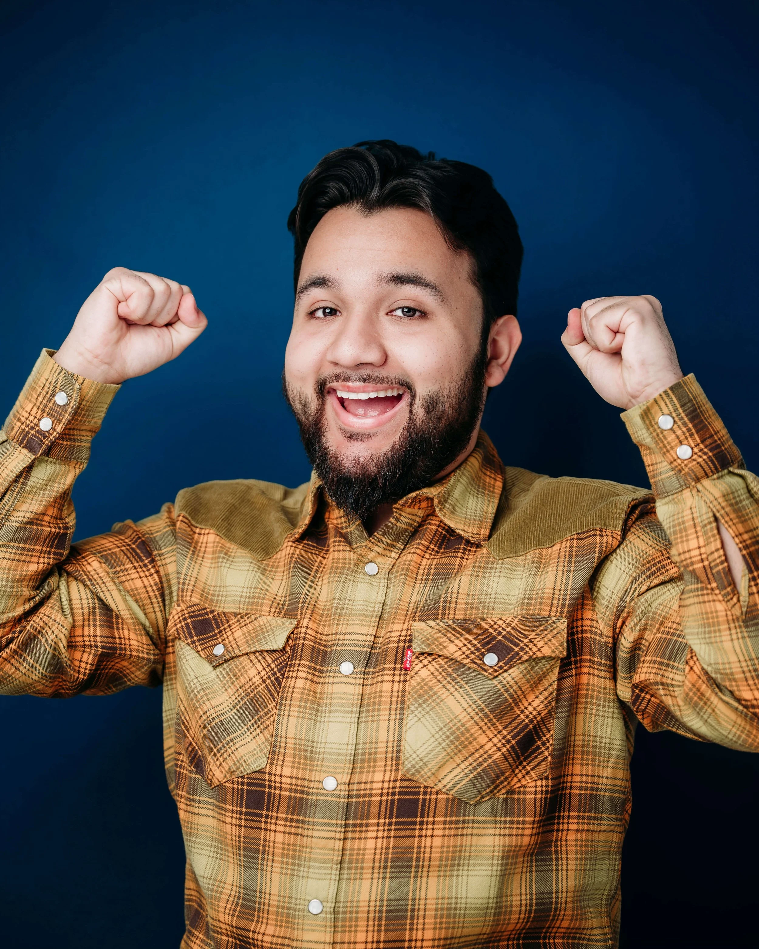 A cheerful man with a beard and dark hair, wearing a mustard plaid shirt, smiling and raising both fists in celebration against a dark blue background.