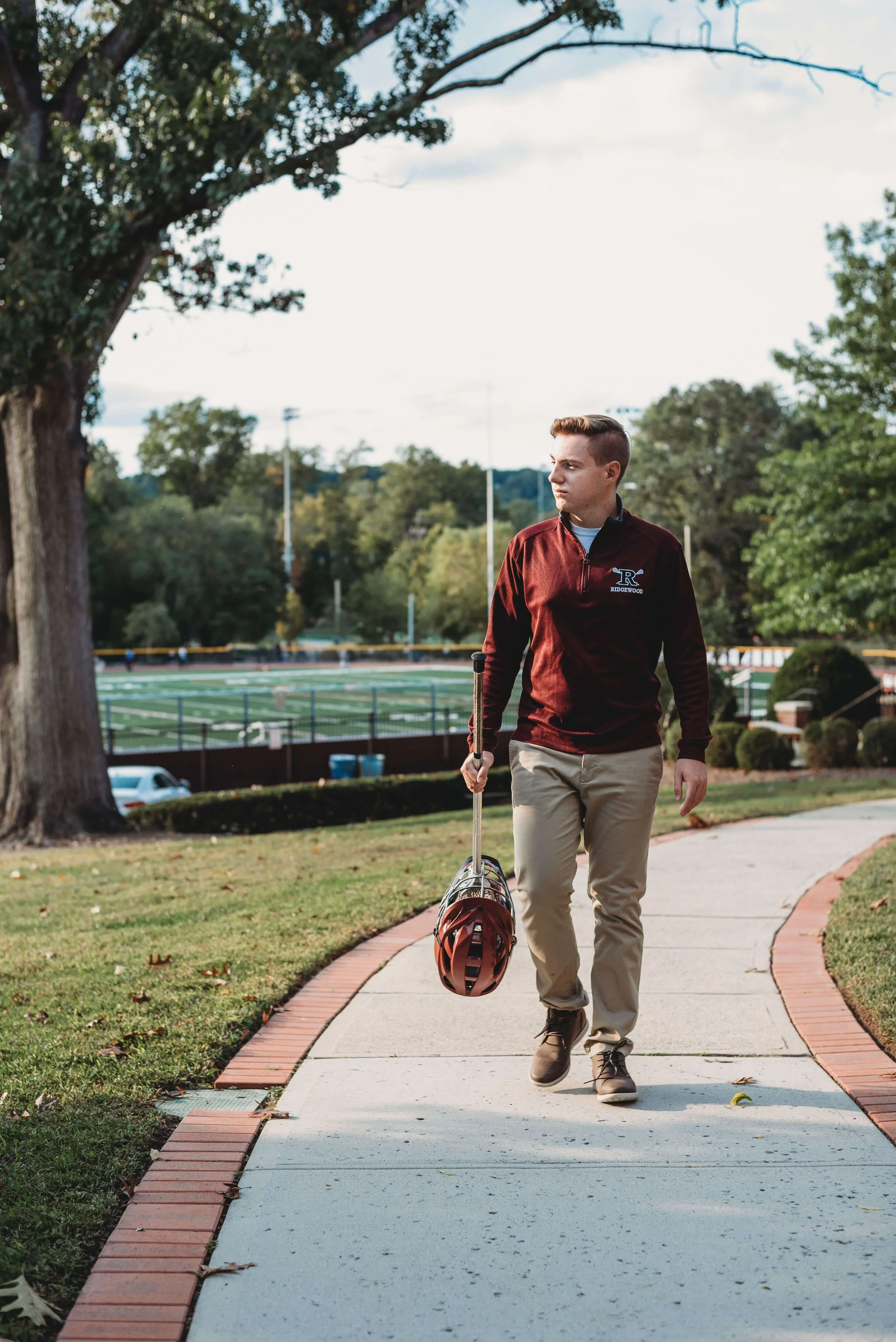 Young man walking on a sidewalk holding a football helmet in his right hand, wearing sports attire with a team logo, in a park near a football field with trees and a cloudy sky in the background.