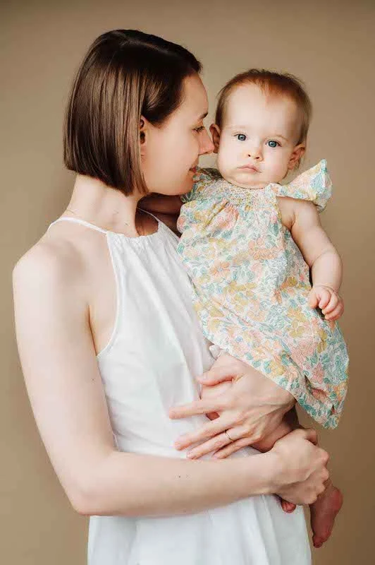 A woman with short brown hair holds a young girl with light hair and blue eyes, wearing a pastel-colored floral dress, against a neutral background.