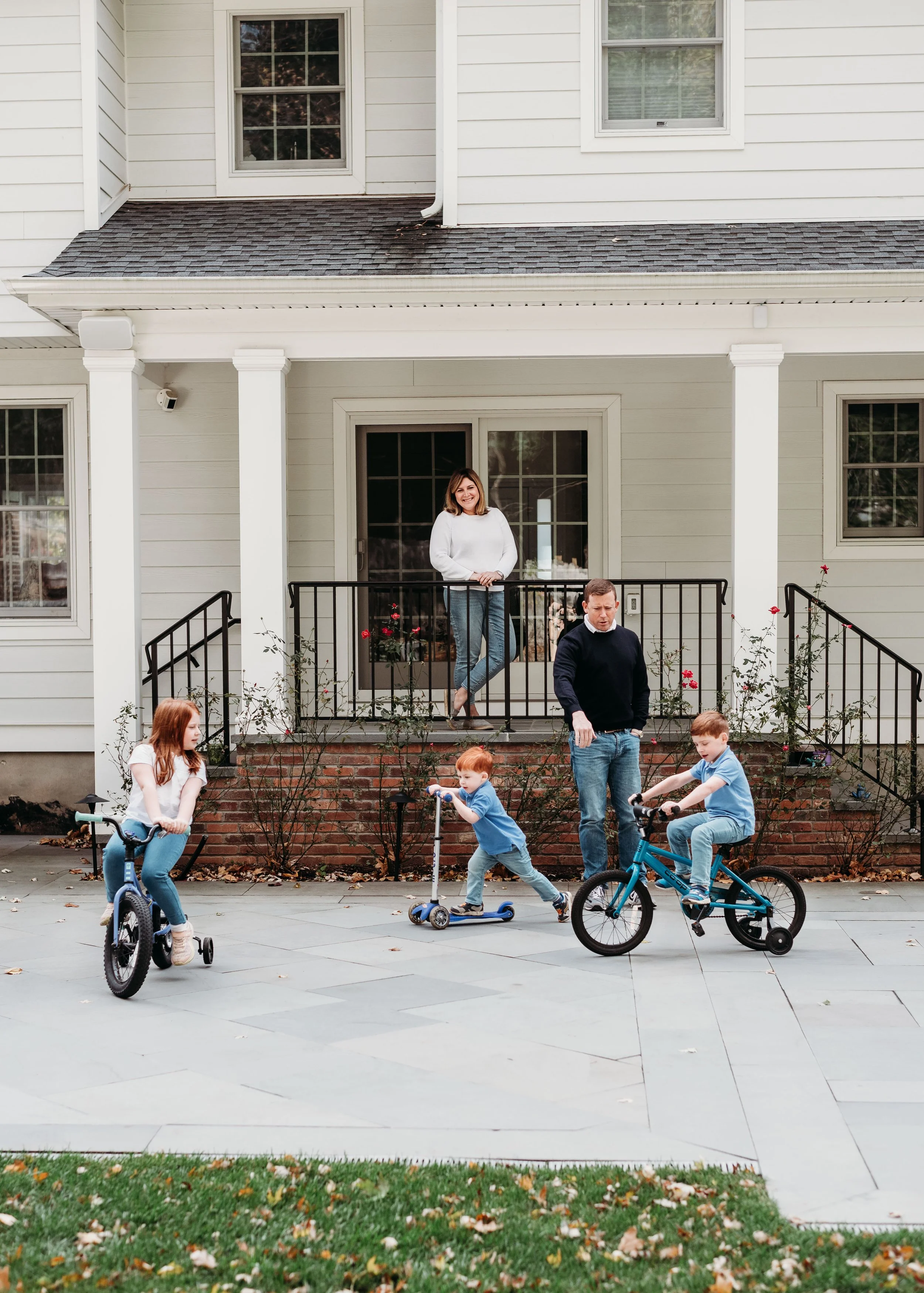 A family enjoying outdoor activities in front of a white house. A woman is standing on the porch, smiling. Three children are riding bikes and scooters on the sidewalk. The house has patio doors, windows, bushes with roses, and a small set of brick s