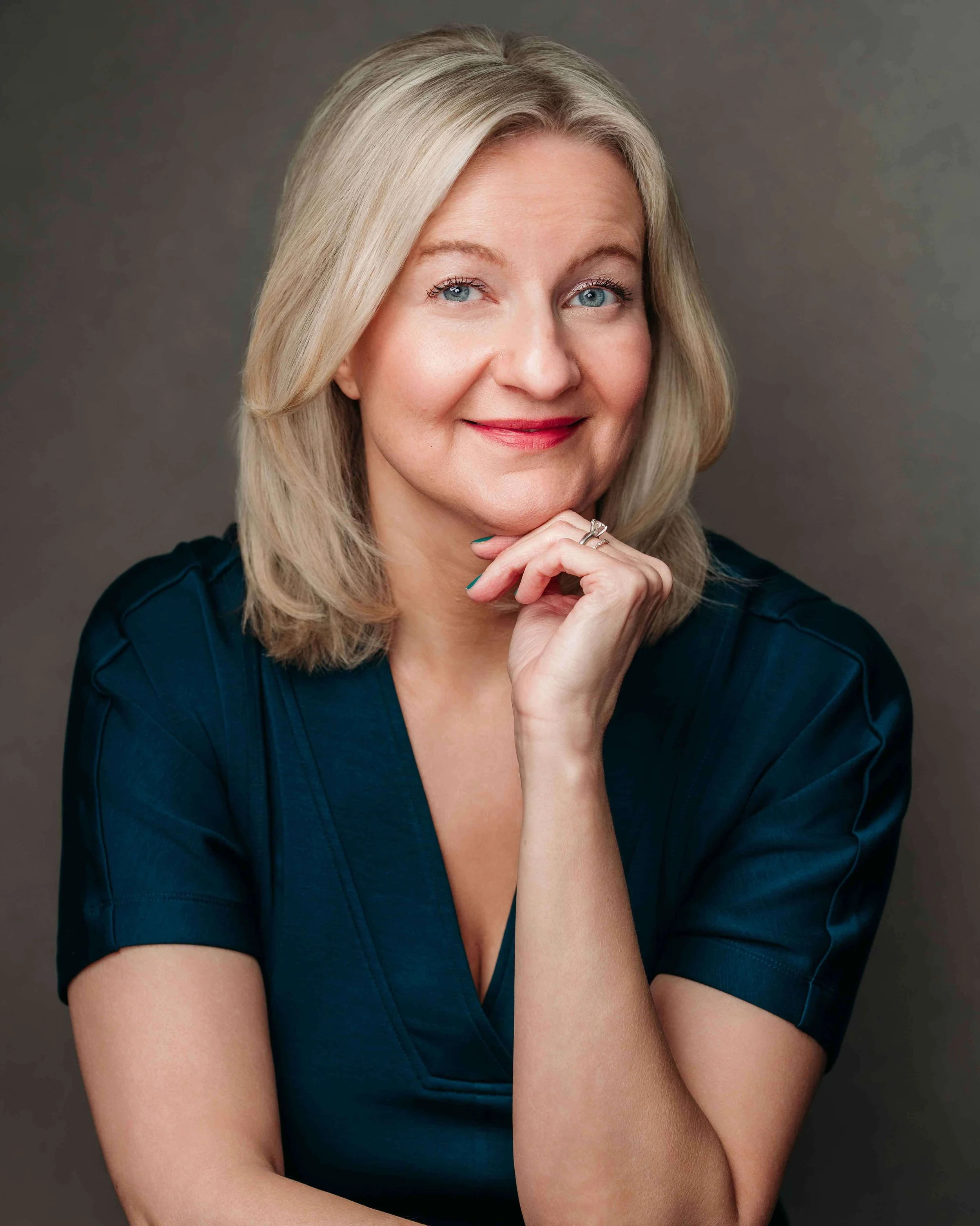 A blonde woman with blue eyes in a blue dress, smiling and resting her chin on her hand against a neutral background.