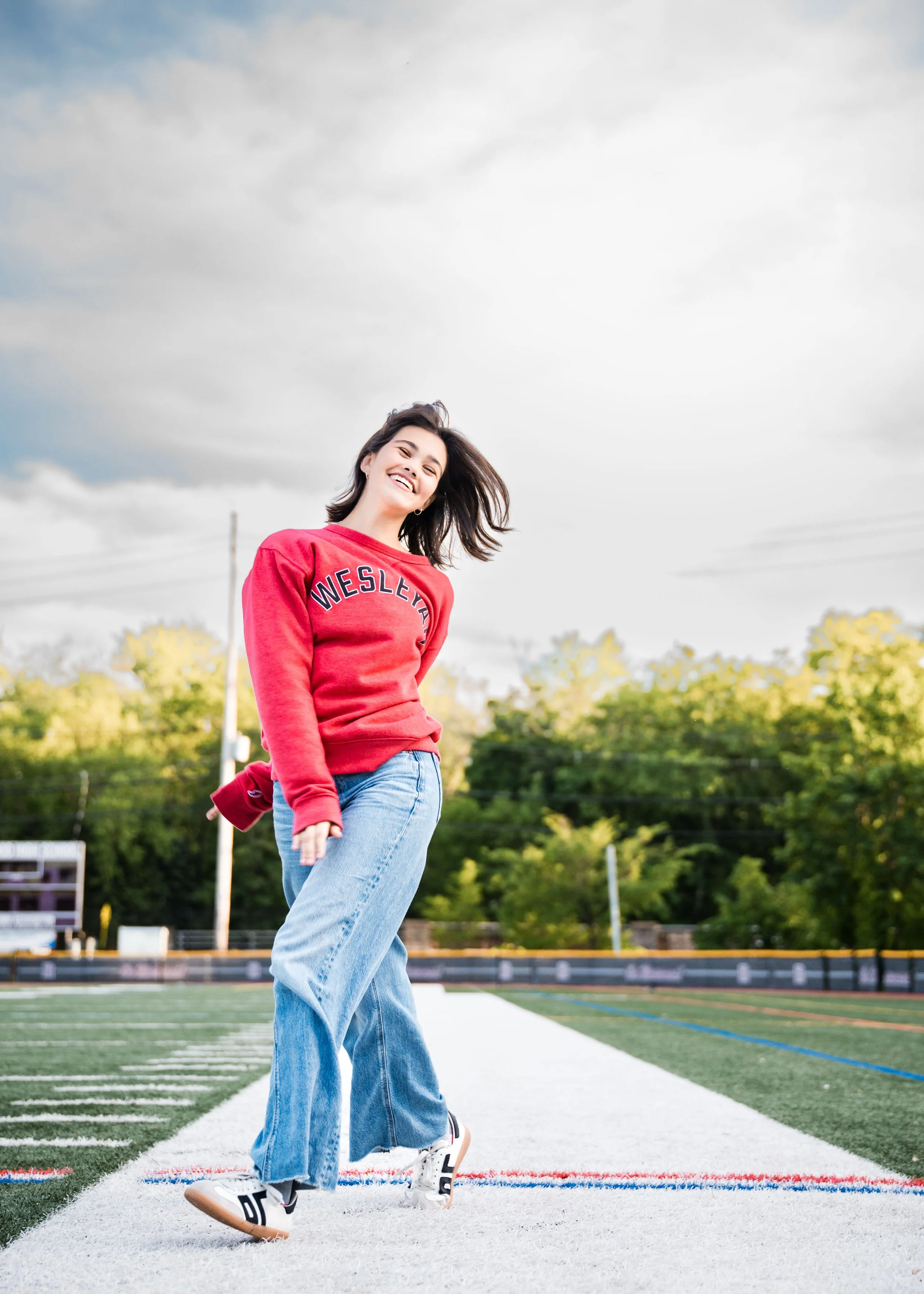 A young woman in a red sweatshirt and blue jeans smiling and dancing on a football field.