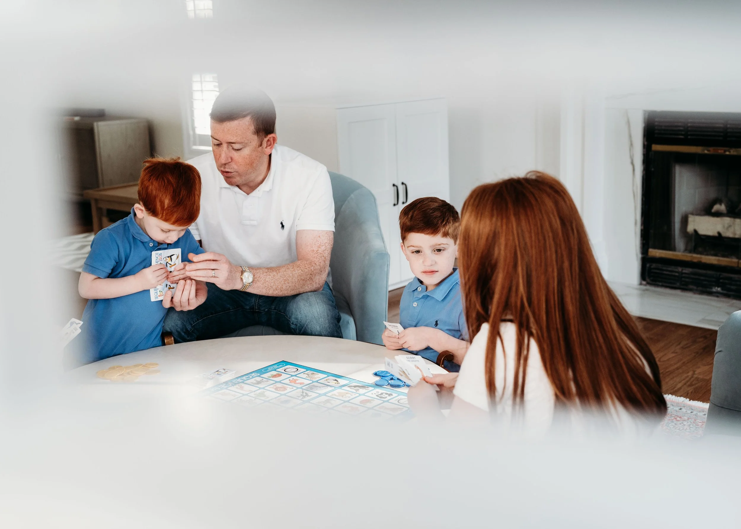 Family playing a board game at a dining table, with two young boys and two adults, in a living room with a fireplace.