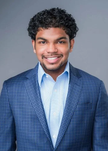 A young man in a blue suit and blue shirt smiling and standing against a light grey background 