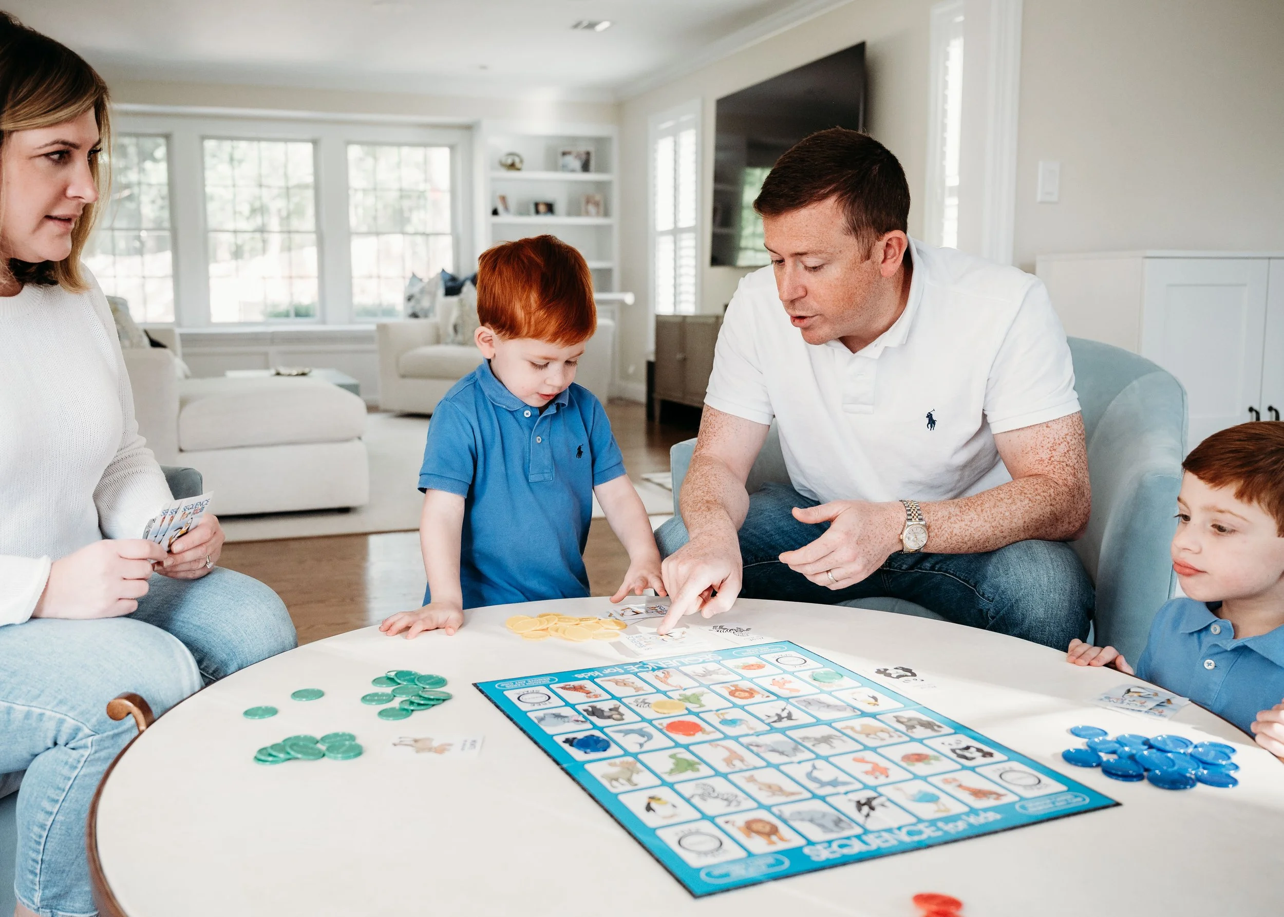 Family playing the board game 'Dixit' around a table in a bright living room with large windows.
