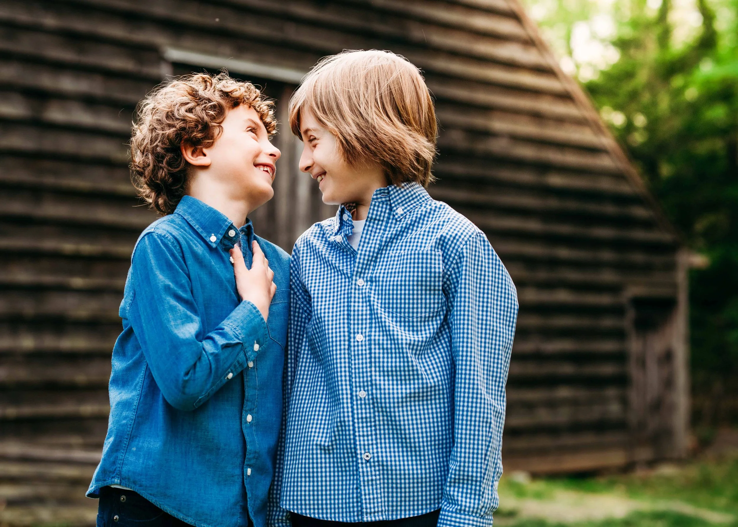 Two young boys face each other smiling, outdoors in front of a wooden barn