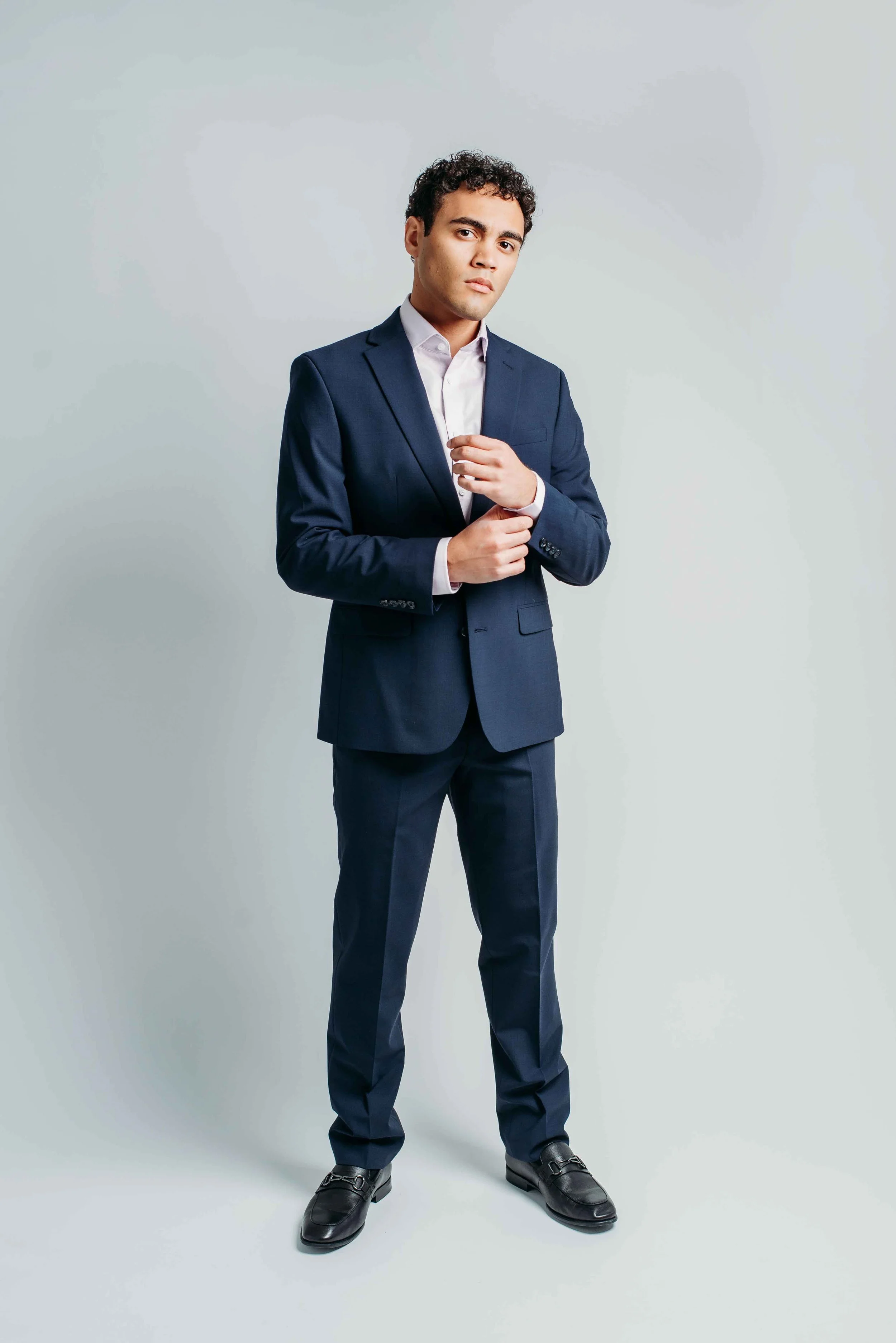 A man in a navy suit adjusting his cuff, standing against a plain light gray background.