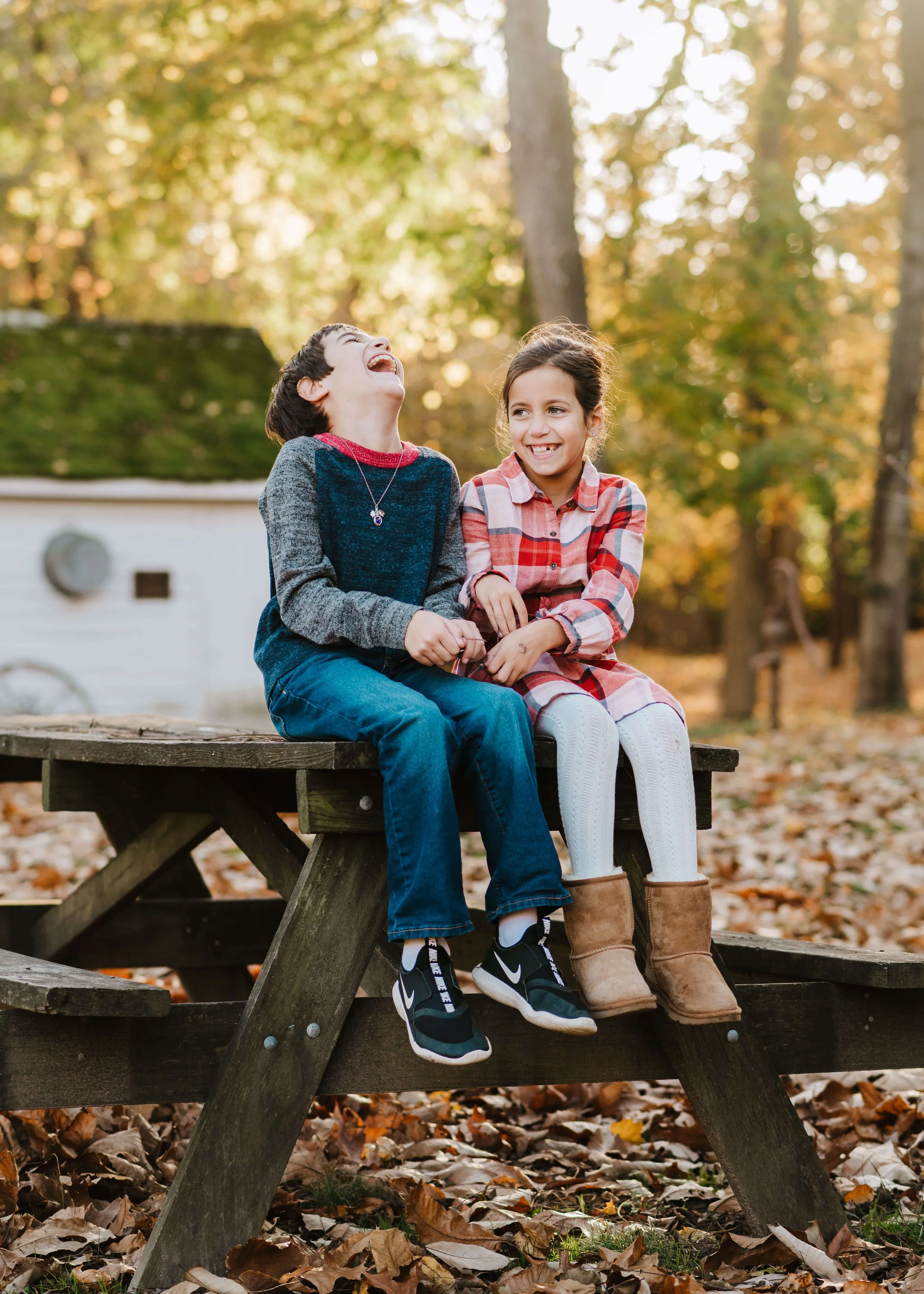 Two children, a boy and a girl, sitting on a wooden picnic table outdoors surrounded by autumn foliage, laughing and smiling.