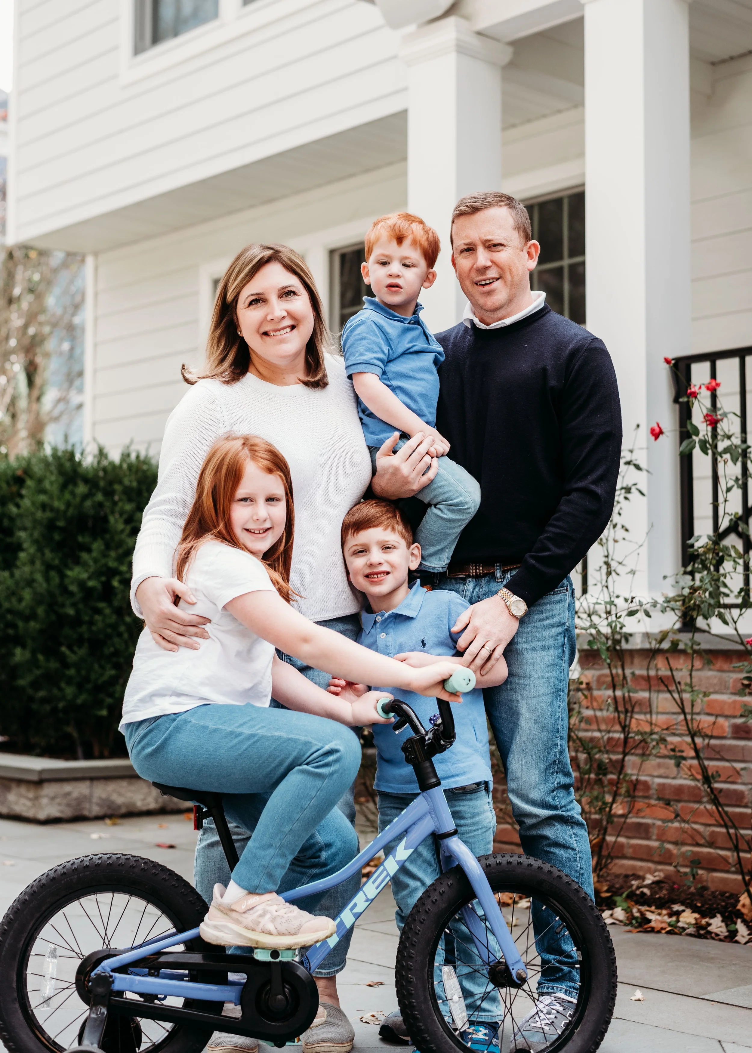 Family of six standing outdoors in front of a house, with a girl on a bicycle, two boys, a woman, and a man smiling at the camera.