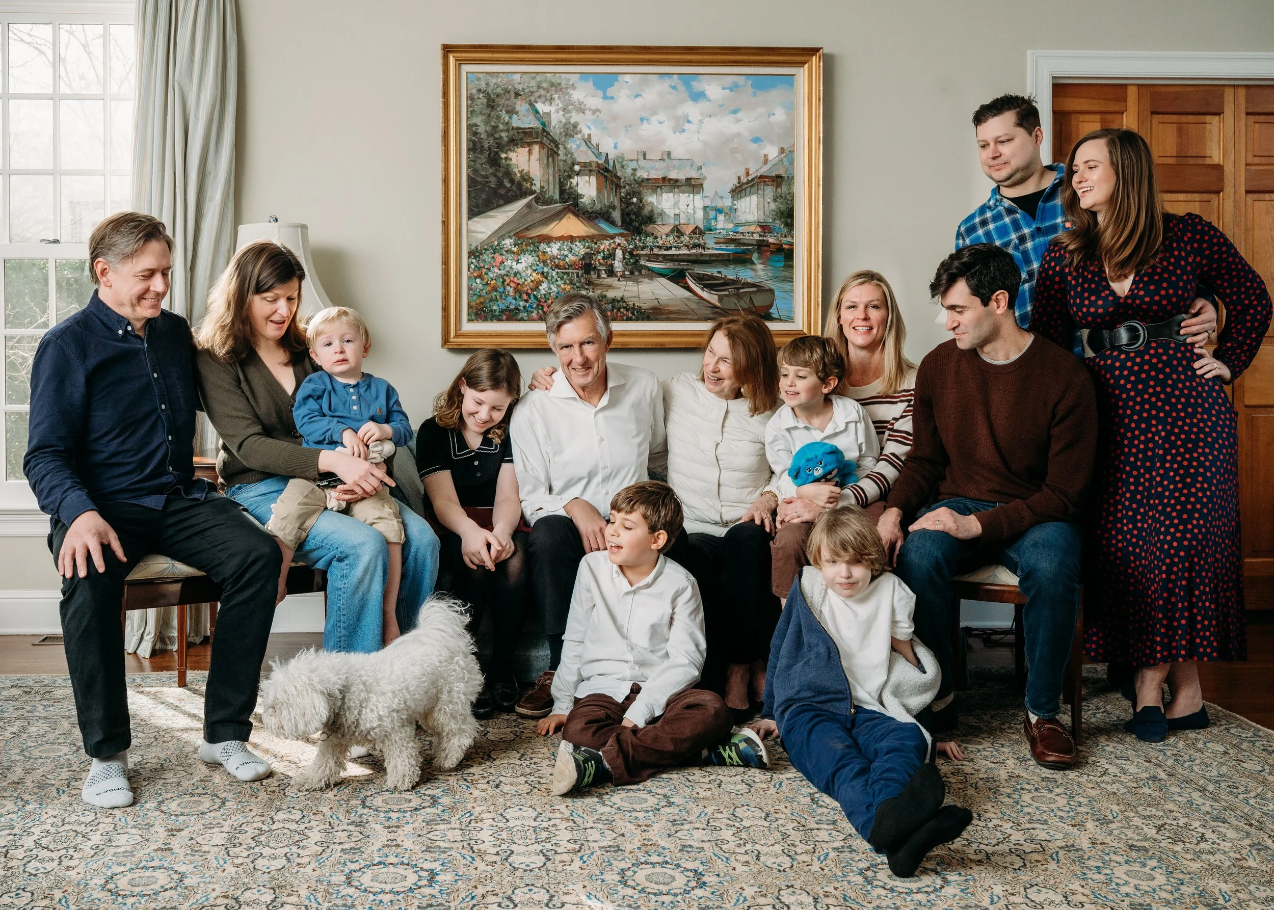 Family gathering in a living room with multiple generations, children, adults, and a pet dog, all sitting and standing around, smiling and engaging with each other, with a large framed painting of a marina scene on the wall behind them.