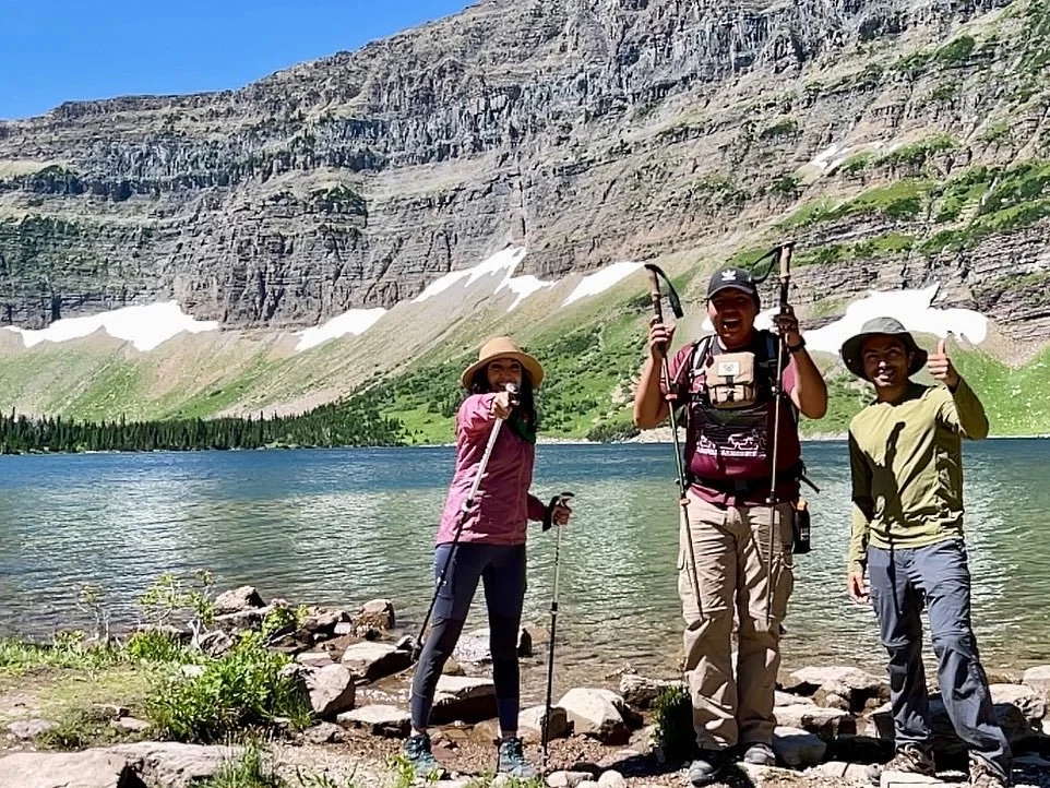 Three people hiking near a lake in a mountainous area, with rocky cliffs and patches of snow in the background, wearing outdoor gear and smiling.