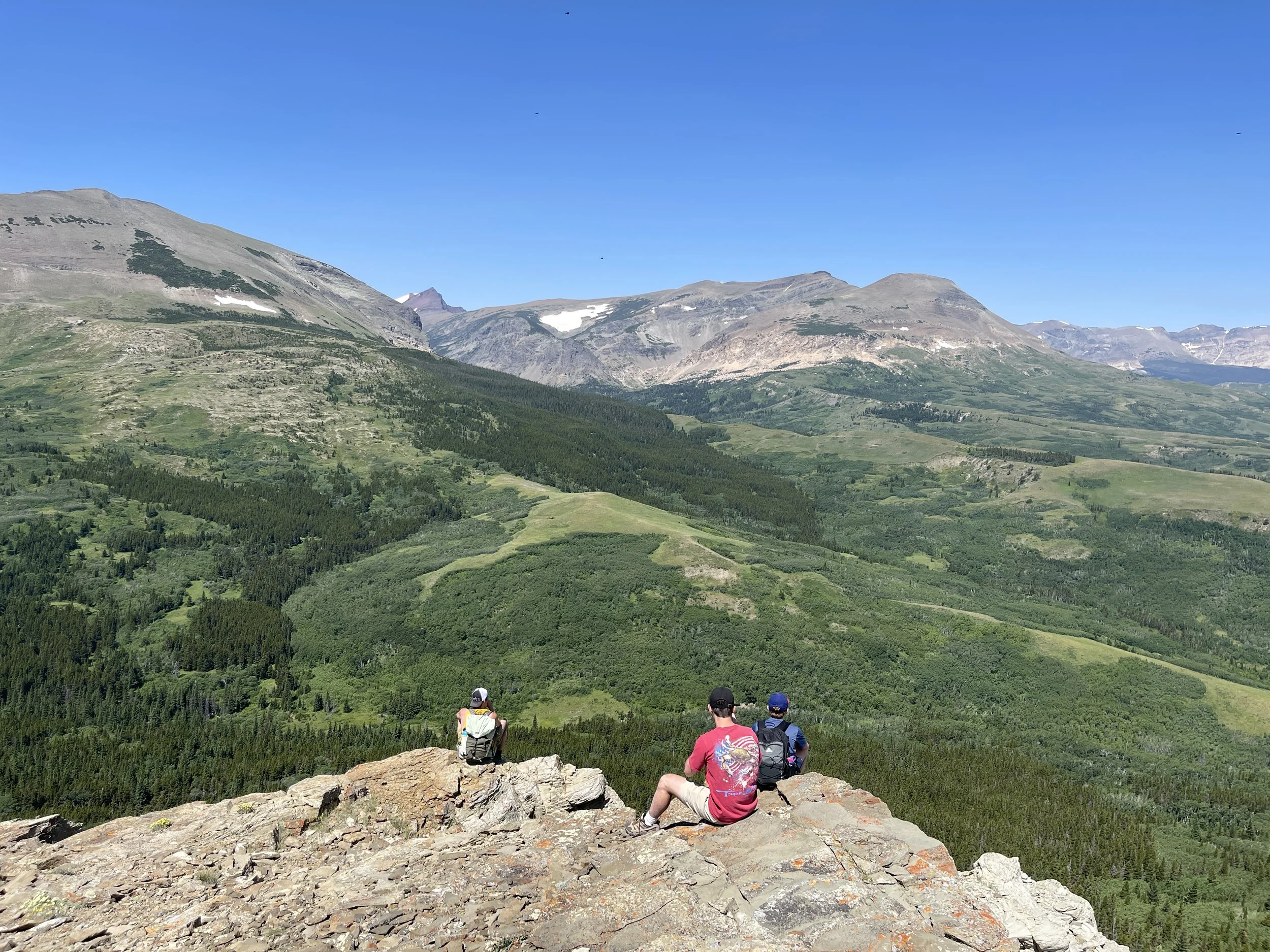 Three hikers sitting on a rocky ledge overlooking a lush green valley with mountains in the background under a clear blue sky.