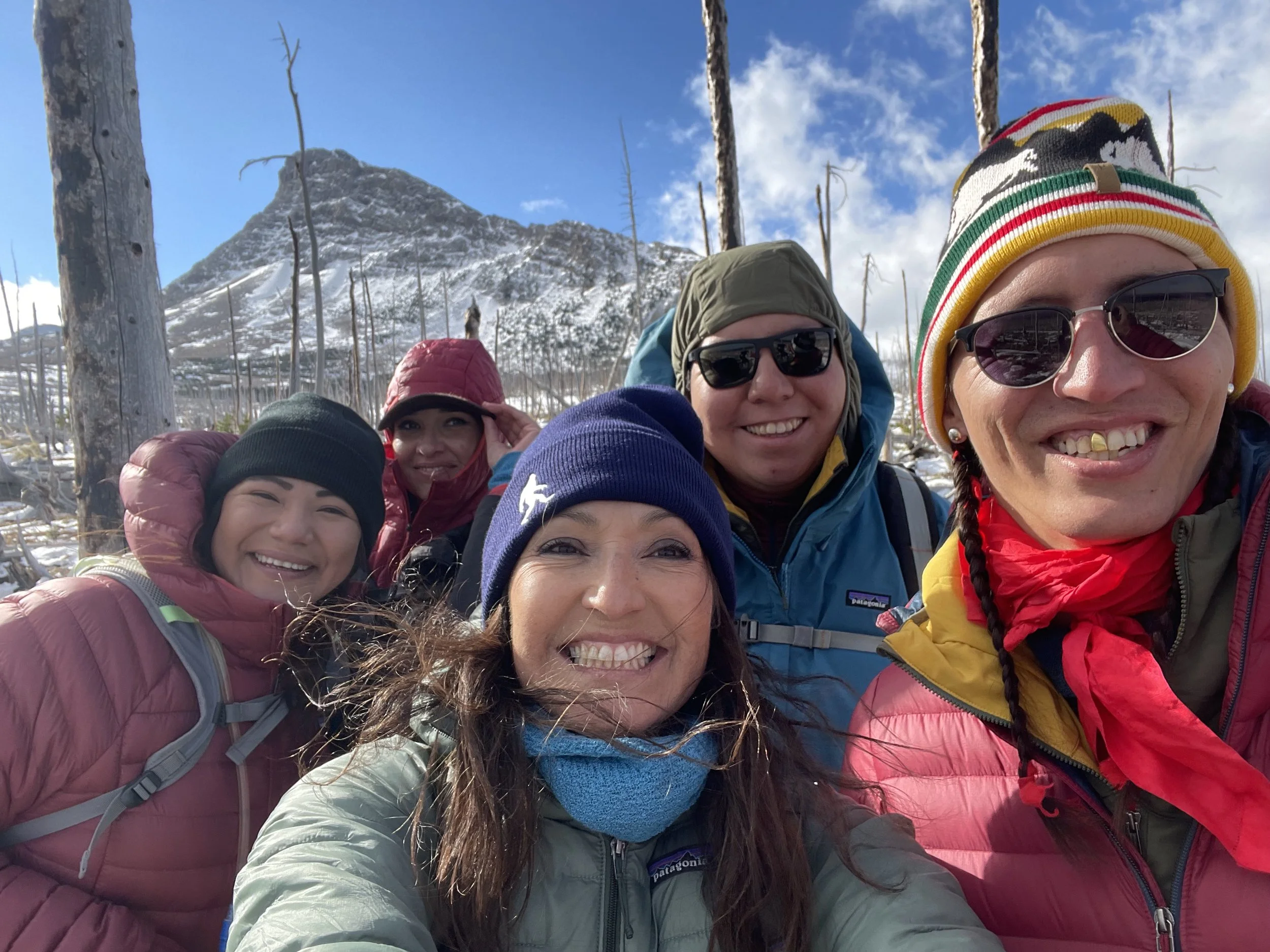 Group of smiling hikers wearing winter gear, sunglasses, and beanies, standing outdoors with snow-covered mountains and a clear blue sky in the background.