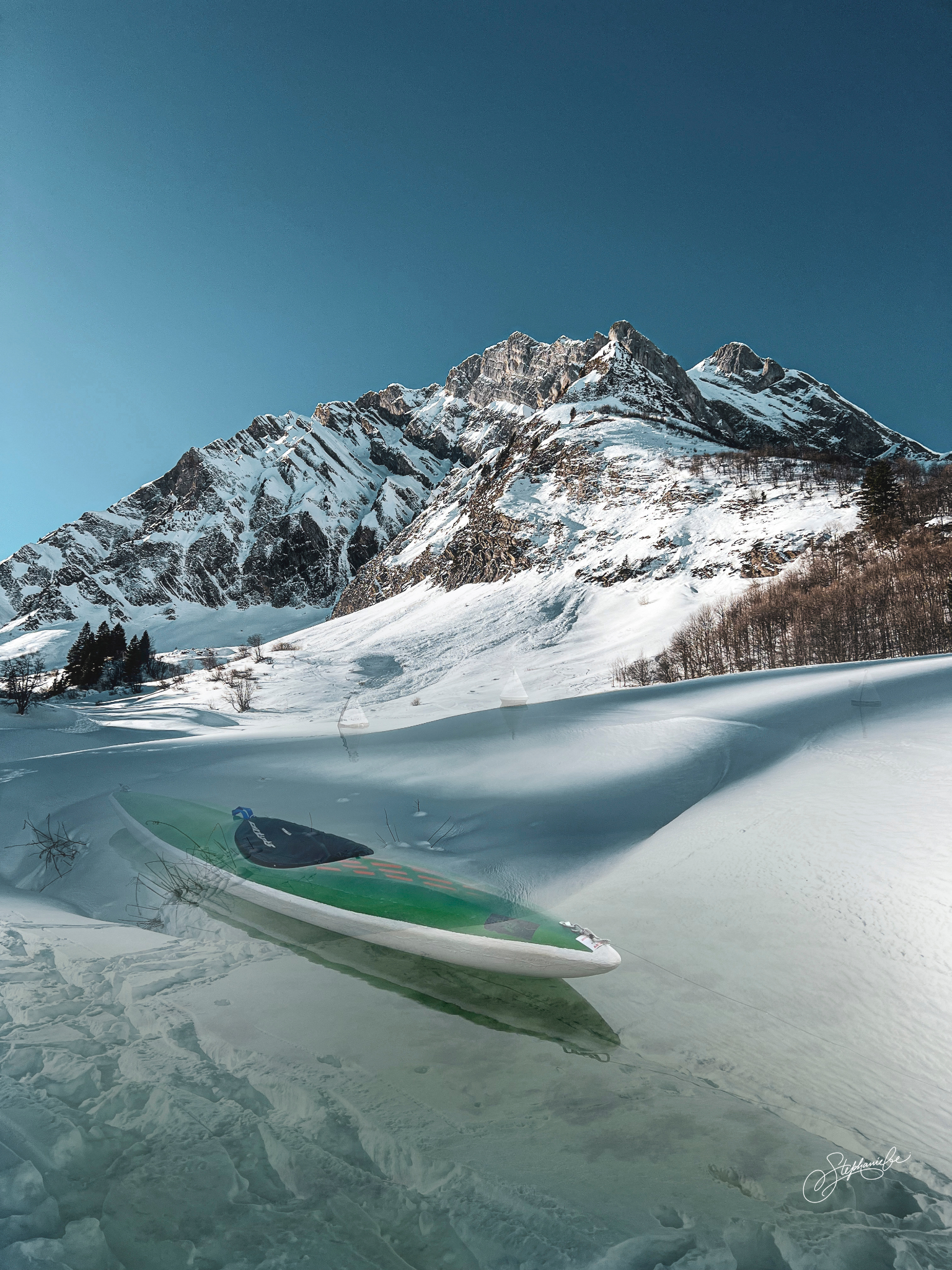 A paddleboard resting on snow-covered ground in front of snowy mountains with clear blue sky.