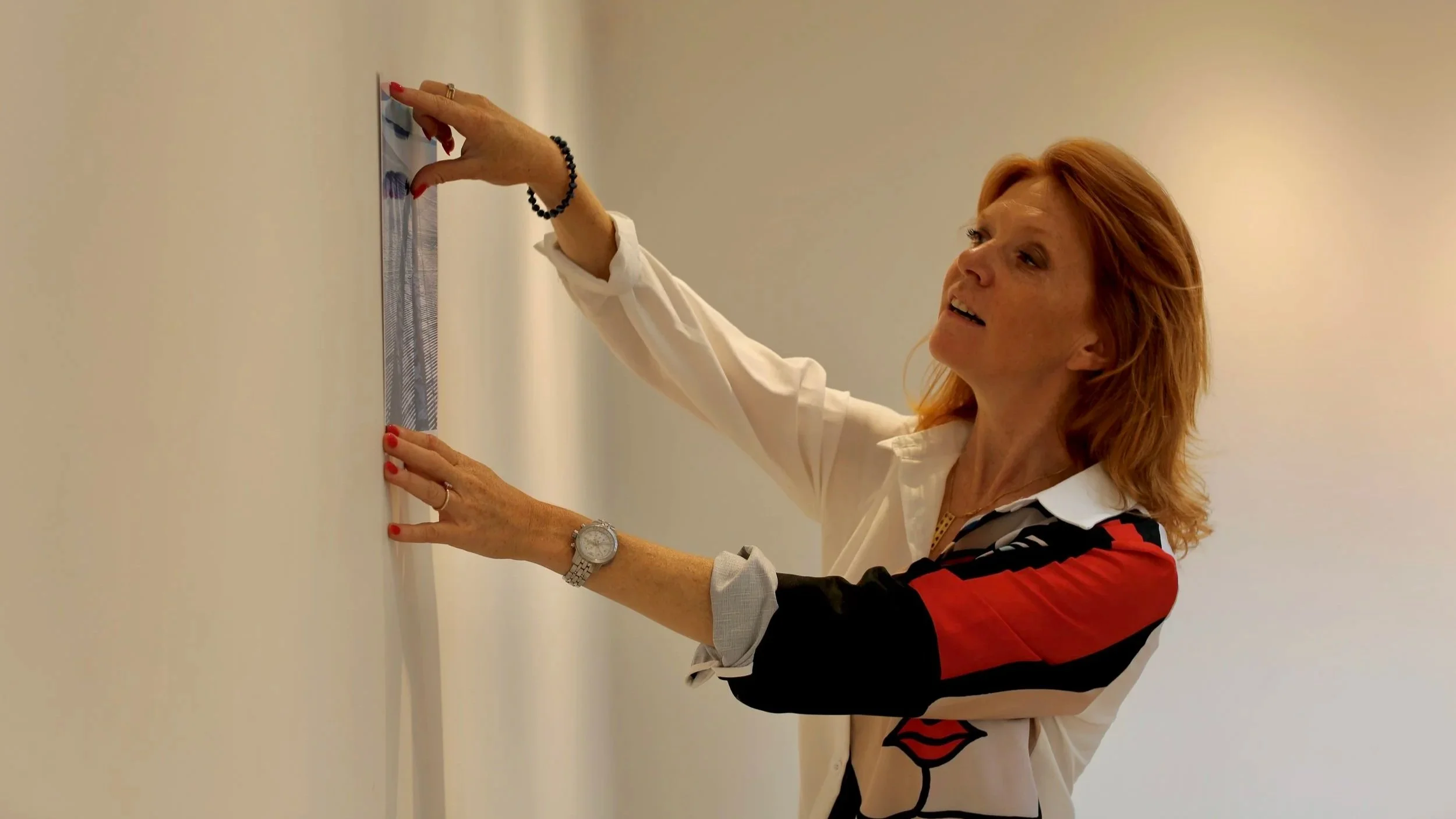 A woman with red hair, wearing a white blouse is measuring a section of a white exhibition wall. She is smiling slightly and has a watch on her left wrist and a black beaded bracelet on her right wrist.