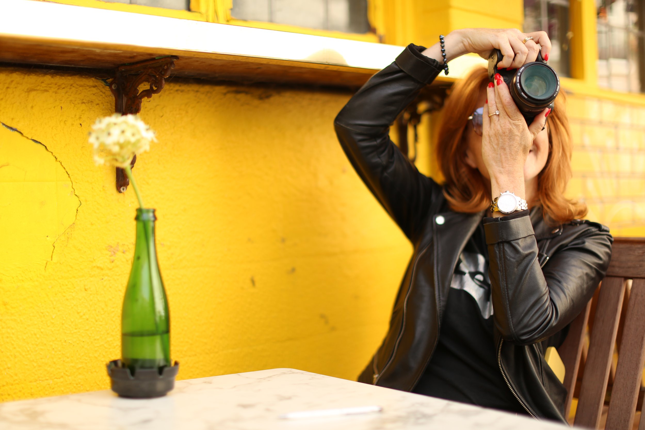 A woman taking a photo with a camera, sitting outside against a yellow wall with a green bottle and a white flower on a table nearby.