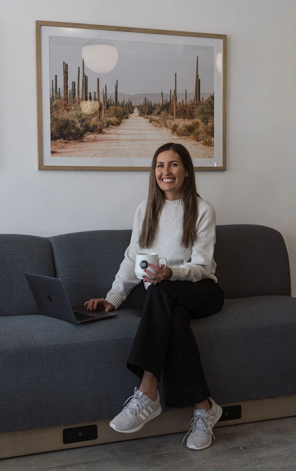 A woman sitting on a gray couch, holding a white mug, with an open laptop on her lap. She is wearing a white sweater, black pants, and sneakers, and is smiling at the camera. Behind her, there is a framed picture of a desert landscape with cacti.