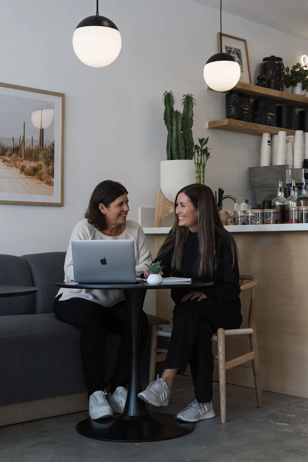 Two women sitting at a small round table in a coffee shop, smiling and talking, with a laptop and a small plant on the table, modern decor with plants and framed art on the wall.