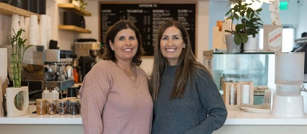 about-picture-Two smiling women standing behind a counter in a coffee shop or cafe with various kitchen items and a menu board in the background.